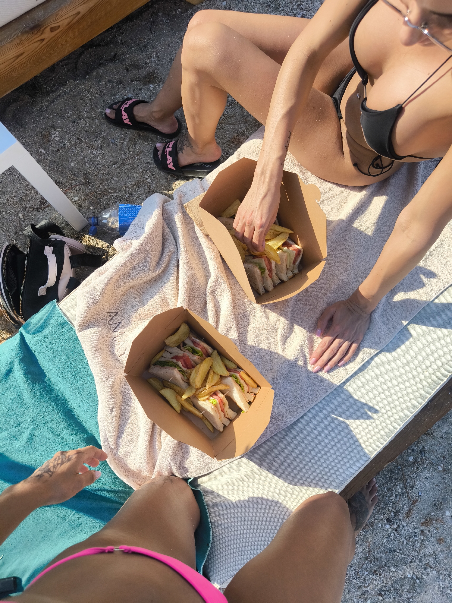 Two women are relaxing on a beach with food. The woman on the left is wearing black flip-flops with pink writing that says "PLAY". She has a tattoo on her left calf. The woman on the right is wearing a black bikini and has a tattoo on her left arm and a piercing on her chest. Both women have their legs visible. In front of them are two cardboard containers filled with sandwiches and fries. They are sitting on a light-colored towel, which is placed on a lounge chair. A turquoise towel is visible to the left of the light-colored towel, and a black and pink bag is next to it. A plastic water bottle is also visible on the sand. The lighting suggests it is daytime, likely sunny. The scene appears to be casual and relaxed, possibly a picnic or a beach day.