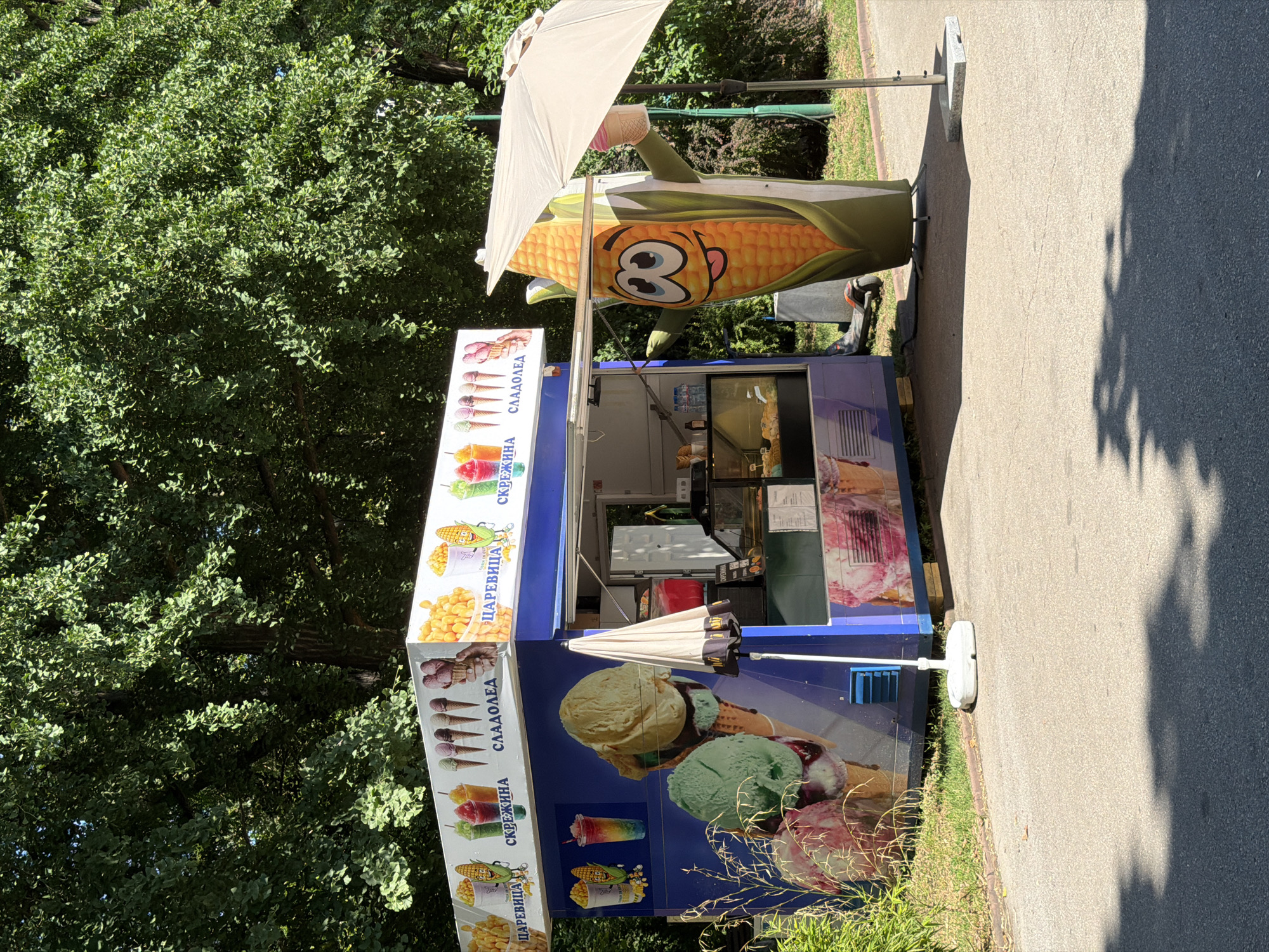 This is a full shot of an ice cream and corn stand located outdoors, likely in a park or public walkway in Pazardzhik, Bulgaria. A large, cartoonish mascot of a corn cob, with a friendly expression and wearing an umbrella, stands to the right of the stand. The stand itself is blue with colorful advertisements for ice cream cones, frozen drinks, and corn. The ground in front of the stand is a paved pathway, and behind it, lush green trees provide shade. A portion of a large tree is visible in the upper background. There are no people visible in the immediate vicinity of the stand.