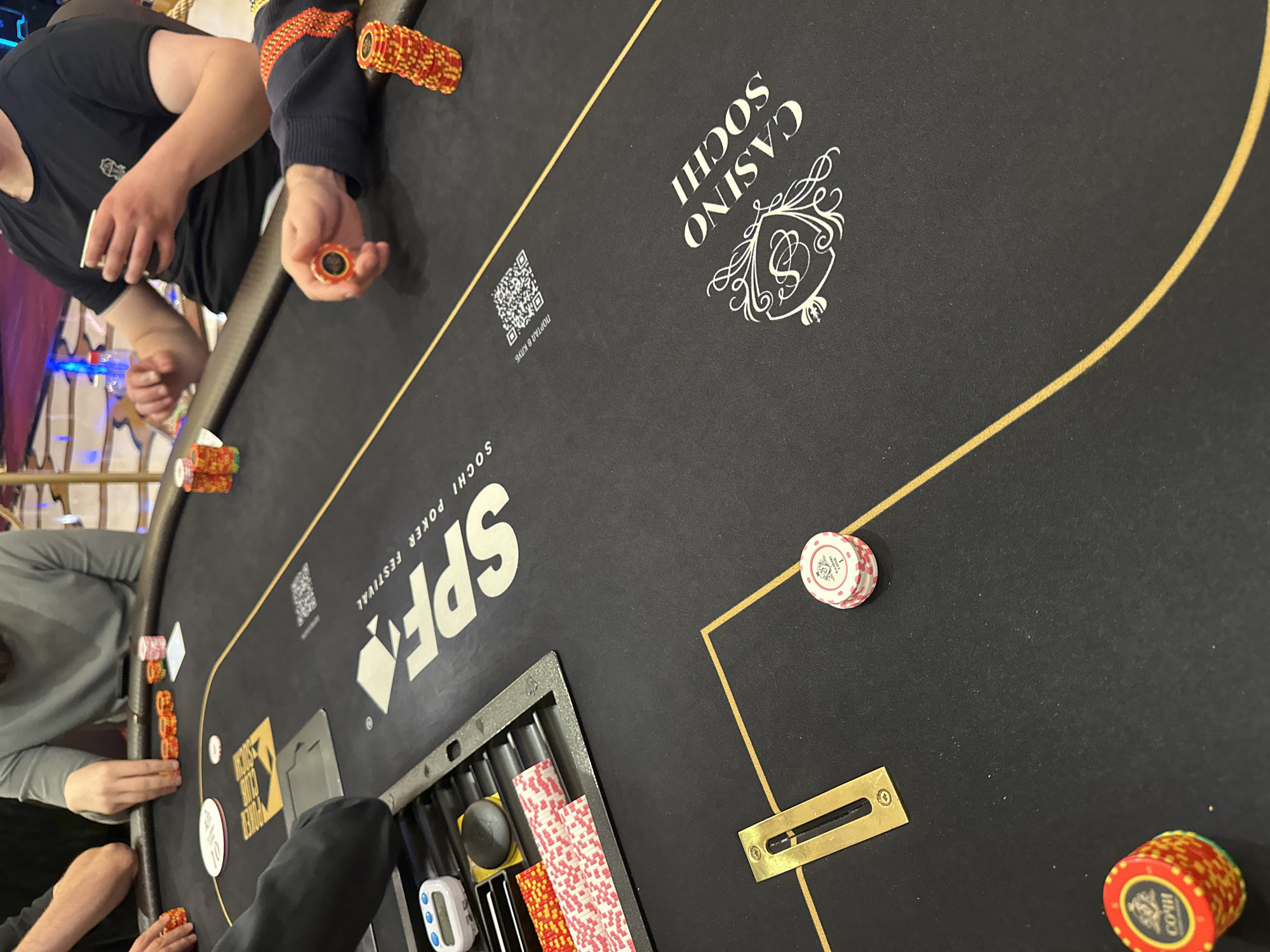 In the city of Estosadok, Russia, a poker game is in progress at the Casino Sochi. The scene is captured from a top-down perspective, focusing on a portion of the black felt poker table. Several stacks of colorful poker chips are visible on the table, indicating betting activity. A player in a gray long-sleeved shirt is seated, with their hands near a stack of orange chips. To their right, another player in a black t-shirt holds a white cup and is also near chips. A third individual, wearing a blue and orange striped sleeve, holds a single orange chip, seemingly engaged in the game. The table features prominent branding, including the "SPF SOCHI POKER FESTIVAL" logo and a QR code, suggesting a tournament or special event. There is also a metal tray with various items, including a small digital timer and slots containing cards and chips. A golden inlay track marks the perimeter of the playing area on the table. The overall environment appears to be indoors, with subtle lighting suggesting a casino setting.