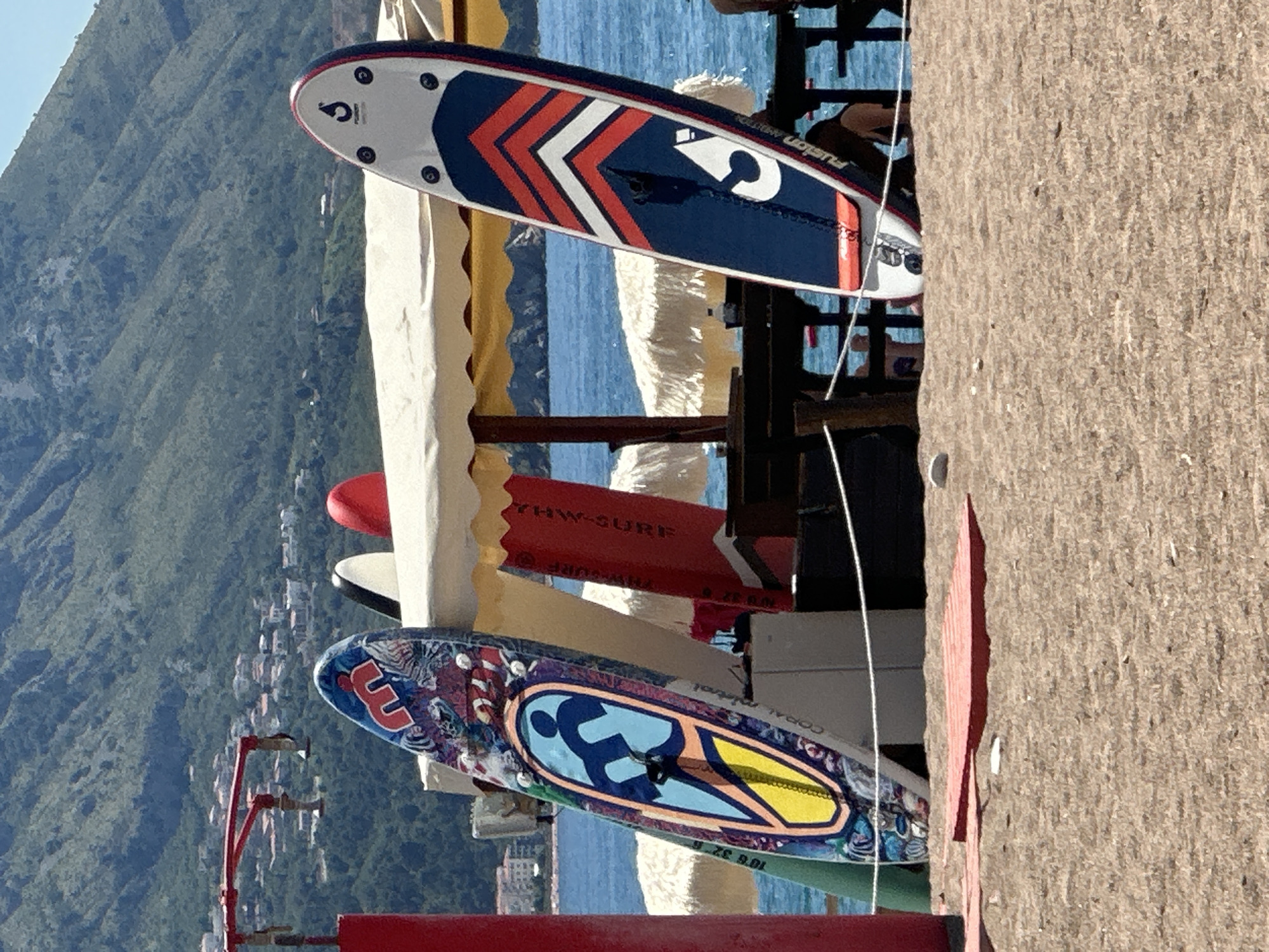 This image depicts a sunny beach scene in Boreti, Montenegro, focused on several paddleboards and surfboards.
In the foreground, light brown sand covers the ground. A white rope barrier stretches horizontally across the lower part of the frame, supported by dark wooden posts. To the left of the rope, two flat, rectangular, slatted red objects rest on the sand.
Dominating the midground are multiple stand-up paddleboards and surfboards leaning against or positioned near beach furniture under a large, light-colored canopy. From left to right:
* A vibrant paddleboard with a multi-colored, abstract design features a central graphic of a person on a board. Text "CORAL MISTRAL" and "10'6 32 6" is visible on its side. It leans against a red pillar which is part of a red metal frame structure on the far left.
* Behind this board, a green board is partially visible.
* Under the canopy, several other boards are present. A red surfboard with the partially visible text "THW-SURF" is prominent. Another white board with a black tip and a red board are also discernible.
* To the right, a dark blue, white, and red paddleboard stands upright, featuring a large white "S" logo and the text "FUSION AMBITION".
The canopy, mostly white with a visible yellow scalloped edge on the right, covers dark wooden tables and chairs. Partially visible figures are seated at these tables on the right side of the image.
Beyond the beach, the calm, blue sea stretches into the distance. Several white, mushroom-shaped beach umbrellas or structures are visible on the water's edge or floating in the mid-distance.
In the background, a large, steep, and heavily vegetated mountain range rises under a clear blue sky. Numerous small buildings with white walls and red or terracotta roofs are scattered across the lower slopes of the mountains.
