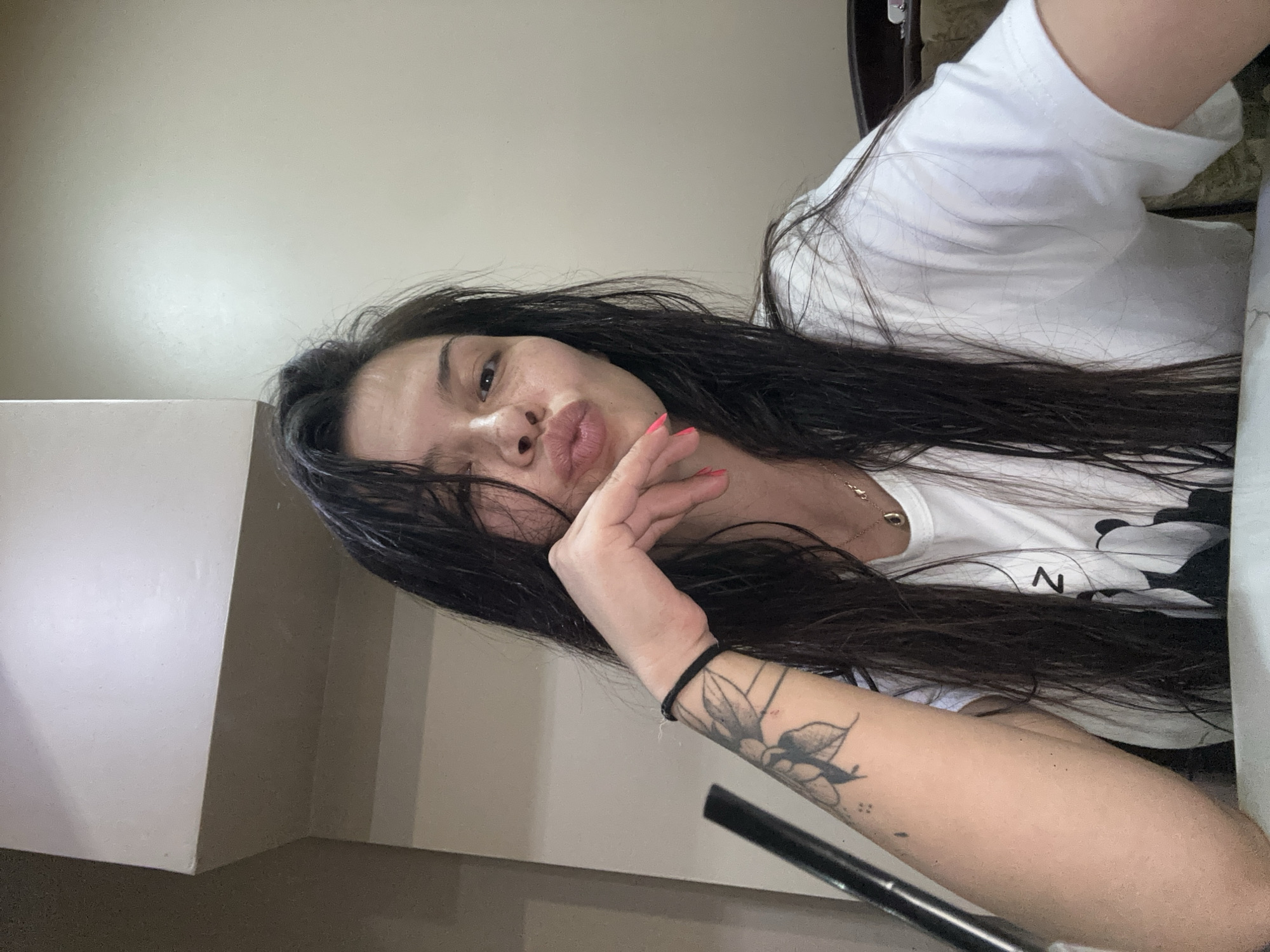 A selfie-style portrait of a woman with long, dark, wet hair, puckering her lips and resting her chin on her hand. She has a tattoo on her left forearm and is wearing a white t-shirt. The background is a plain, light-colored wall with a darker, horizontal element above. The image appears to have been taken indoors. The location in Sliven, Bulgaria is not discernible from the visual information provided.