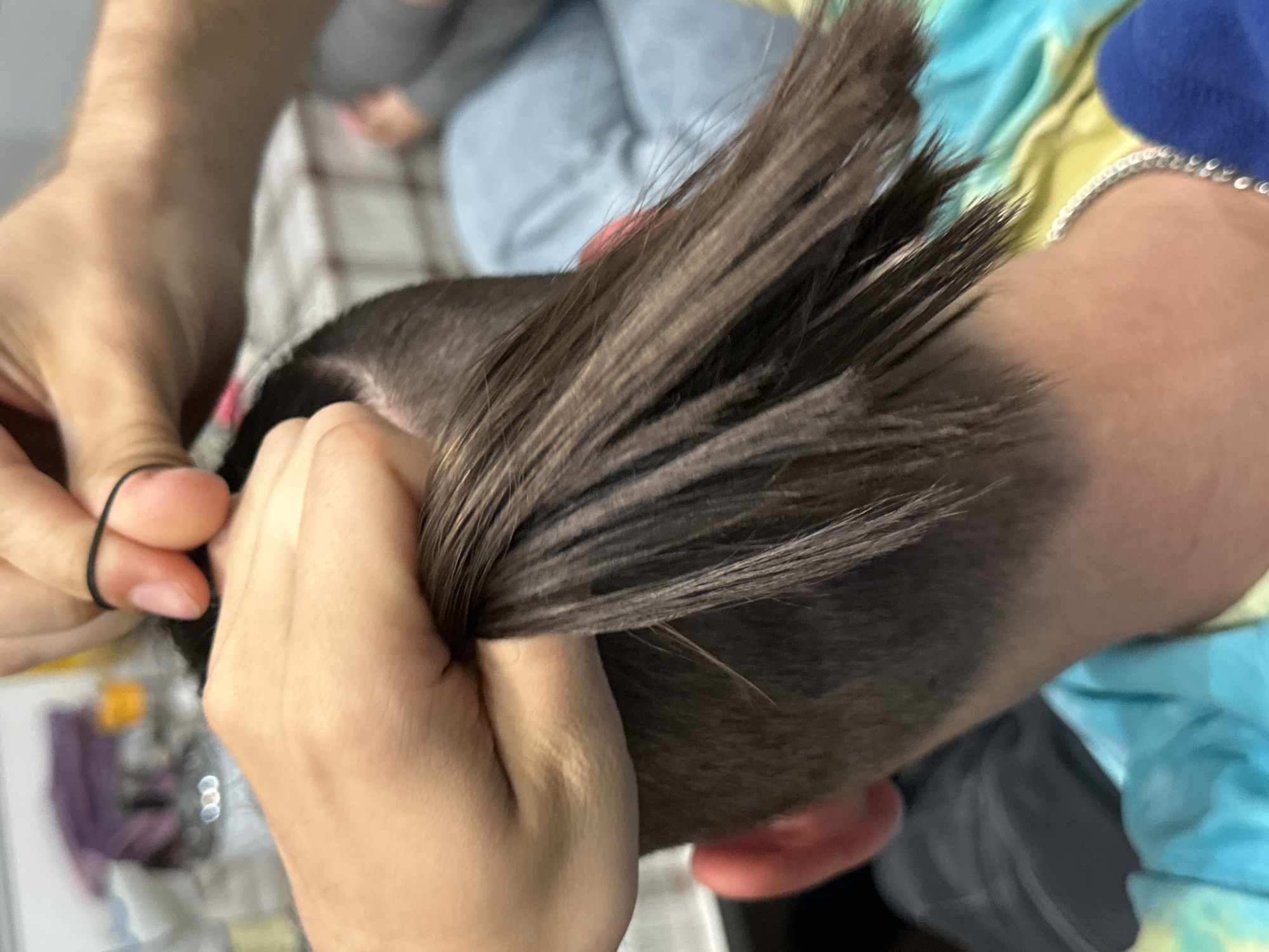 A close-up shot depicts a person's head, with the focus on hands tying the hair up with a black hair tie. The hair is dark brown and styled upwards. The person tying the hair is wearing a colorful shirt, and a silver bracelet is visible on their wrist. Another person's legs are out of focus in the background. The location is unknown and there's no evidence to suggest it's Lovech, Bulgaria.