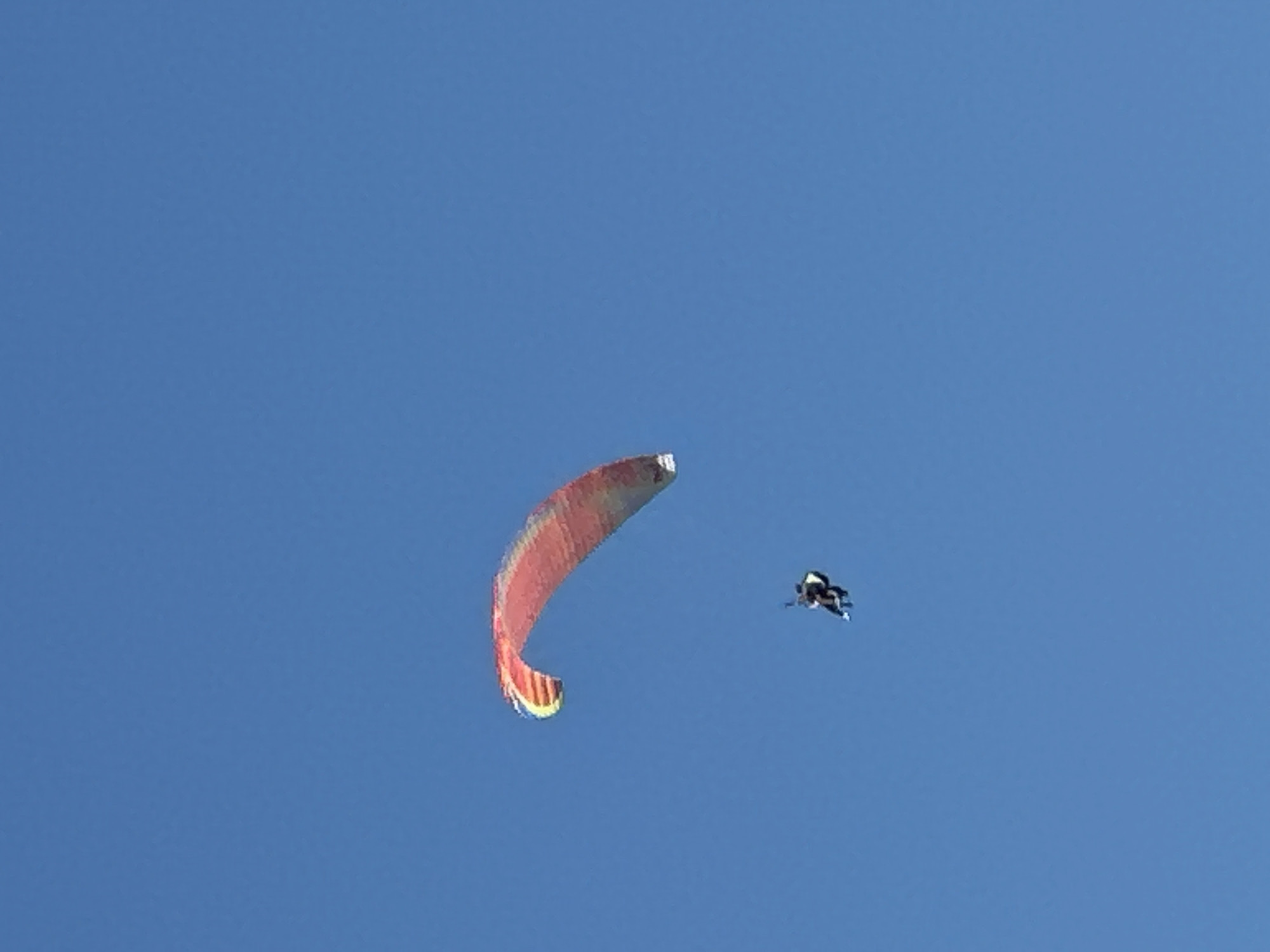 A paraglider, carrying two individuals, is suspended against a clear blue sky. The paraglider canopy is a gradient of red, orange, yellow, and green, with the red section prominently visible in the upper left quadrant of the image. Below and to the right of the paraglider, two figures are seated in a harness. The sky is a uniform, cloudless blue, with no discernible landmarks or city structures visible, indicating this event is occurring in an open, aerial environment, possibly over Boreti, Montenegro, though the specific location cannot be confirmed from the visual data alone.