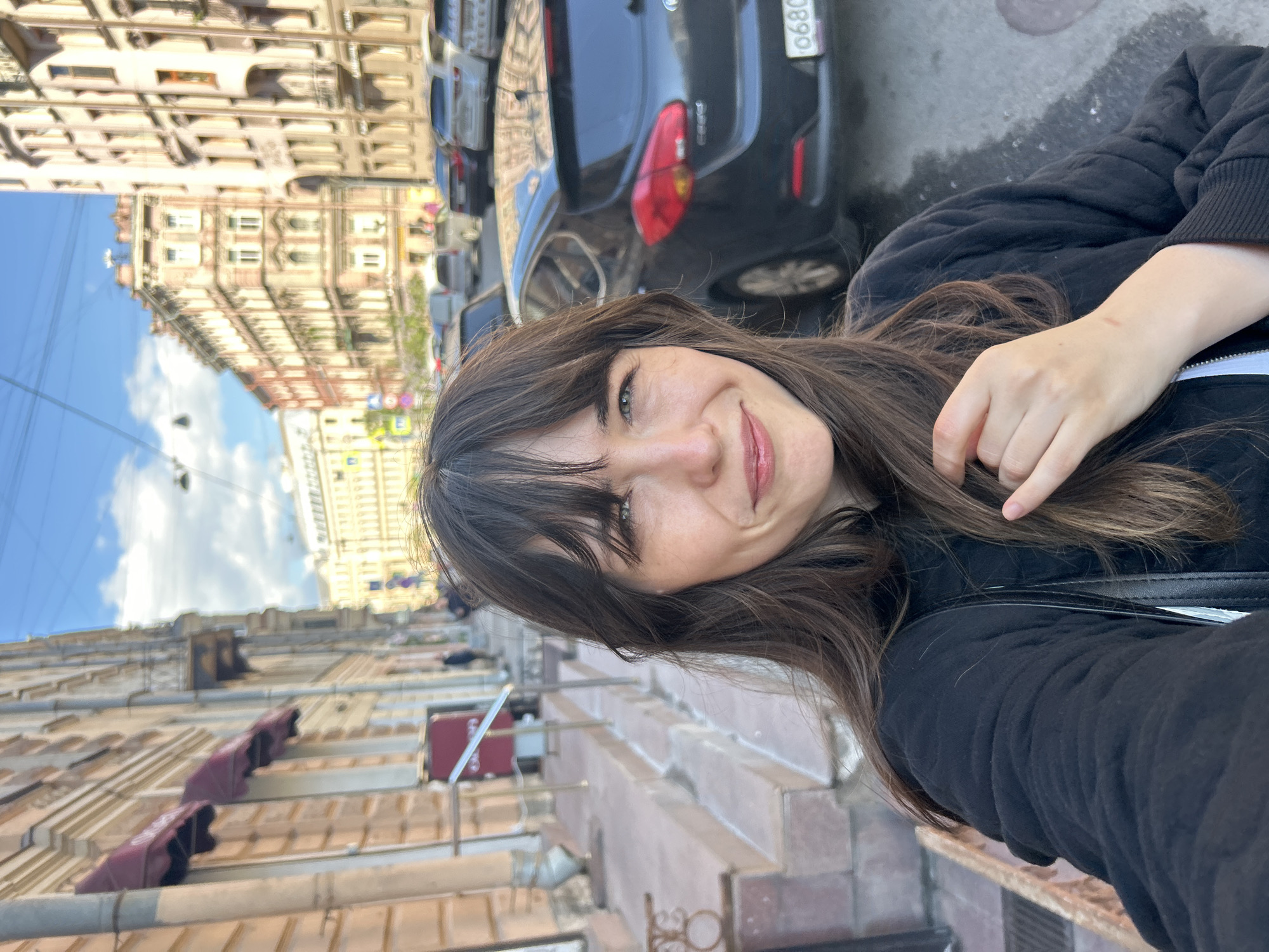 The image is a selfie of a woman with brown hair, a black jacket, and a slight smile, taken on a street in Saint Petersburg, Russia. Behind her, parked cars line the street, with a black car closest to the camera. The architecture is characterized by European-style buildings with detailed facades and awnings. The sky is visible in the upper left corner of the image, with a few clouds. The perspective suggests the photo was taken at street level, likely on the sidewalk.