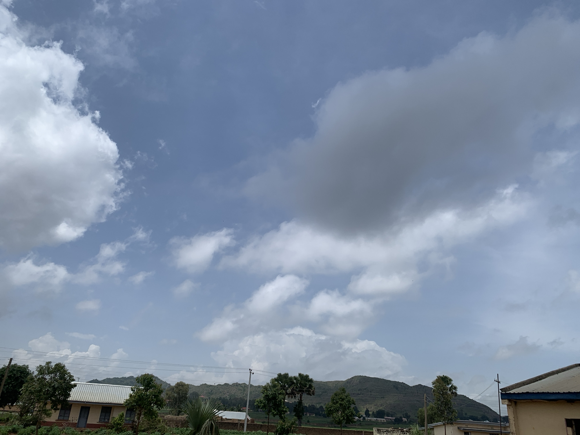 This is a landscape photograph taken in Kuru Karama, Nigeria, under a cloudy sky. The image features a low-lying, multi-story building with a corrugated metal roof in the foreground, partially obscured by various trees. Beyond the building and trees, rolling hills form the backdrop, with more buildings and some vegetation visible at the base of the hills. The sky is predominantly filled with a mix of white and gray clouds, with patches of blue sky visible. The overall scene suggests an outdoor environment with signs of human settlement within a natural landscape.