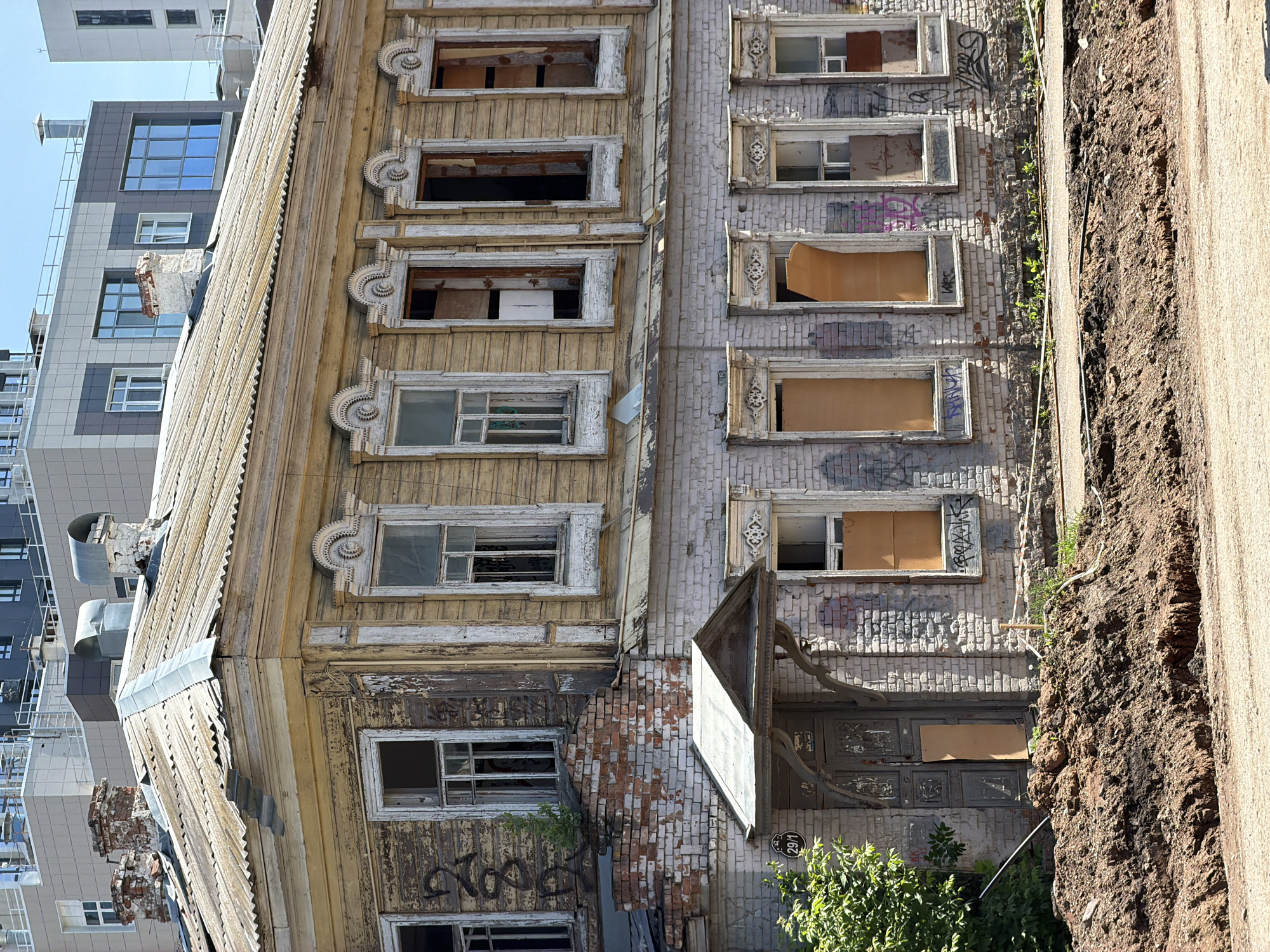 The image shows an old, dilapidated wooden building with a corrugated metal roof in Ufa, Russia. The building is in a state of disrepair, with peeling paint, damaged windows, and graffiti. In the background, modern high-rise buildings are visible, suggesting a contrast between old and new architecture in the city. The foreground shows a dirt pile and road construction, indicating ongoing work or neglect in the area. The scene appears to be an urban environment, likely in a neglected part of the city. No people are visible in the image.
