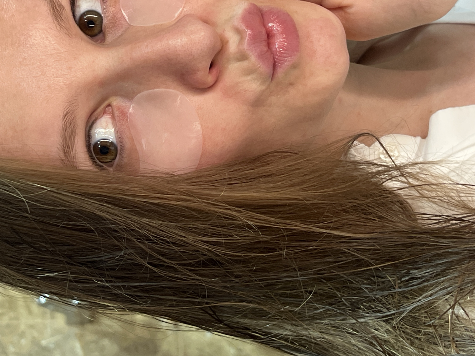A close-up, slightly rotated image shows a woman's face with under-eye gel patches applied. Her eyes are brown, and her lips are slightly pursed and glossy. Long brown hair is visible in the foreground. The background is out of focus and does not provide any recognizable landmarks or features to pinpoint the location as Khimki, Russia.