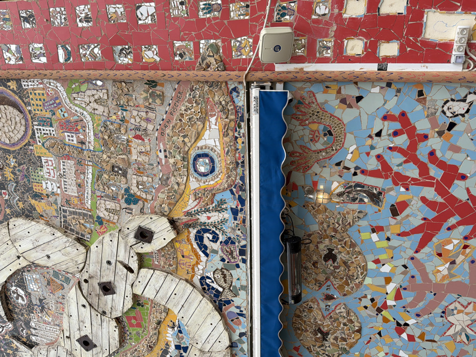 The image displays a detailed view of an exterior building wall covered in elaborate mosaic artwork by Oren Fischer, located at 28 Yehezkel Street, Tel-Aviv-Yafo, Israel, associated with the "Oneg Shabbat" establishment.
The upper and central portions of the primary facade are adorned with a multi-material mosaic. Large, light-colored wooden cable spools with rusted metal square centers are arranged in an abstract, overlapping pattern. Behind and around these, the mosaic depicts a vibrant cityscape with buildings, trees, and abstract figures, utilizing various colored tiles, pebbles, and found objects. The artist's name, "OREN FISCHER," is visible within this section. Below the spool elements, a prominent mosaic eye with a blue and white concentric iris transitions into undulating wave-like patterns and small fish motifs.
Below this upper artwork, a blue fabric awning with a scalloped edge extends horizontally. The awning's valance features Hebrew text: "28 יחזקאל" (Yehezkel 28) and "עונג שבת" (Oneg Shabbat). Beneath the awning, a lower mosaic panel depicts an underwater scene with fish formed from pebbles and tiles, vibrant red coral-like structures, and other abstract marine elements on a light blue background. An elongated black infrared outdoor heater is mounted horizontally on this panel, alongside a single wall-mounted, caged light fixture.
To the right, a narrower vertical wall section is entirely covered in a red mosaic composed of small, irregularly shaped tiles. This red mosaic features multiple recessed square and rectangular frames, each containing miniature mosaic depictions of abstract figures, faces, and patterns. A light-colored square outdoor speaker or sensor with a dark circular emblem is mounted on this red wall. A white electrical conduit runs vertically beneath it, leading to two stacked white double-gang electrical outlets near the bottom. A vertical metal pole or conduit defines the boundary between the main mosaic facade and the red mosaic section. No individuals or dynamic actions are present in the scene.