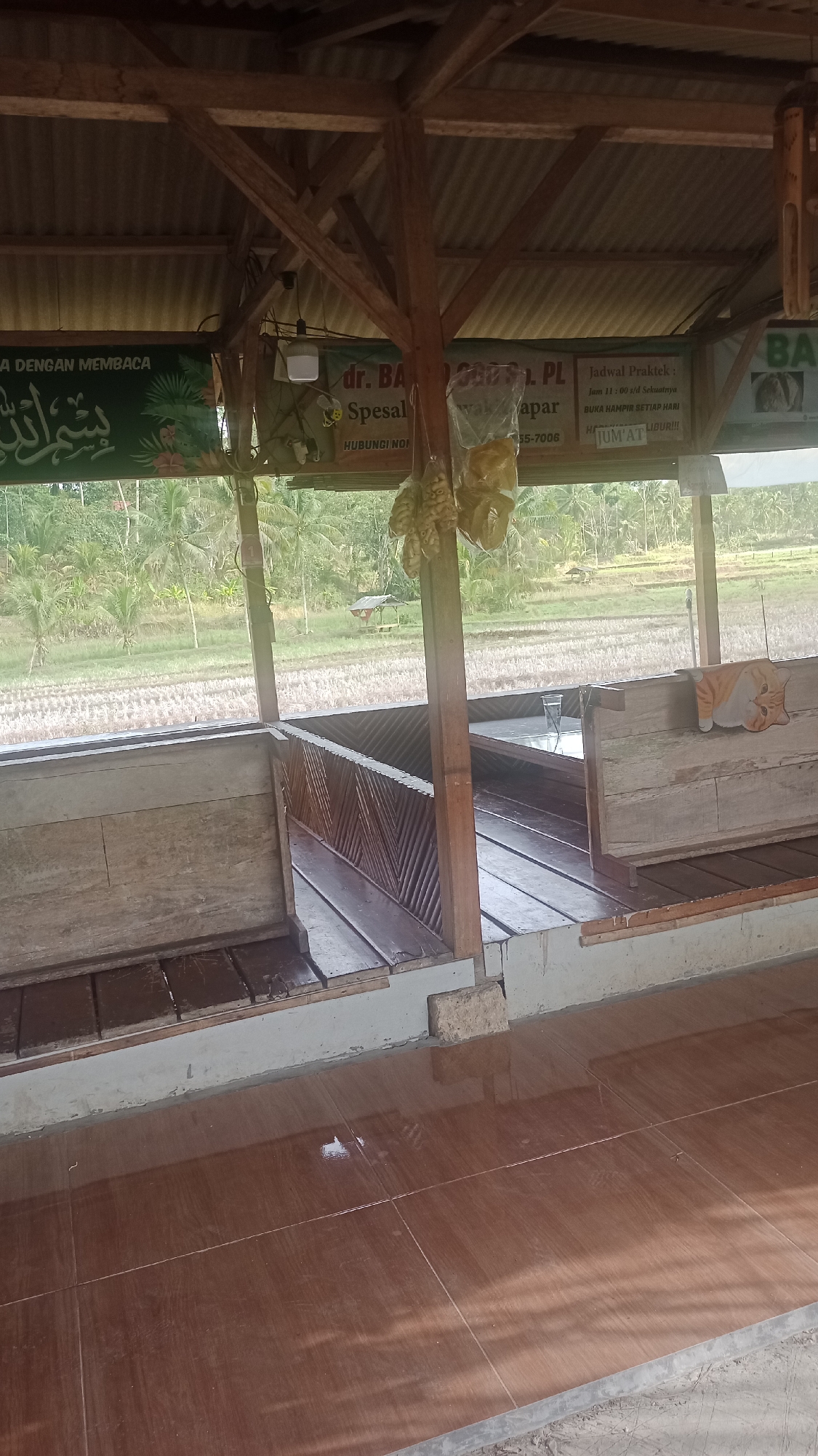 The image depicts an outdoor seating area, likely a warung or small restaurant, in Cibeber, Indonesia. The structure is made of wood and corrugated metal roofing, with open sides offering a view of a rice field and palm trees. A sign is visible with Arabic script and text that appears to be in Indonesian, possibly advertising services or hours of operation. The seating consists of wooden benches and tables. A glass of water sits on one of the tables, and an orange and white cat-shaped cushion is on a bench. The floor is tiled and appears to be wet in places. Hanging from the ceiling are dried food items, possibly snacks. The overall impression is of a casual, rustic establishment in a rural setting.