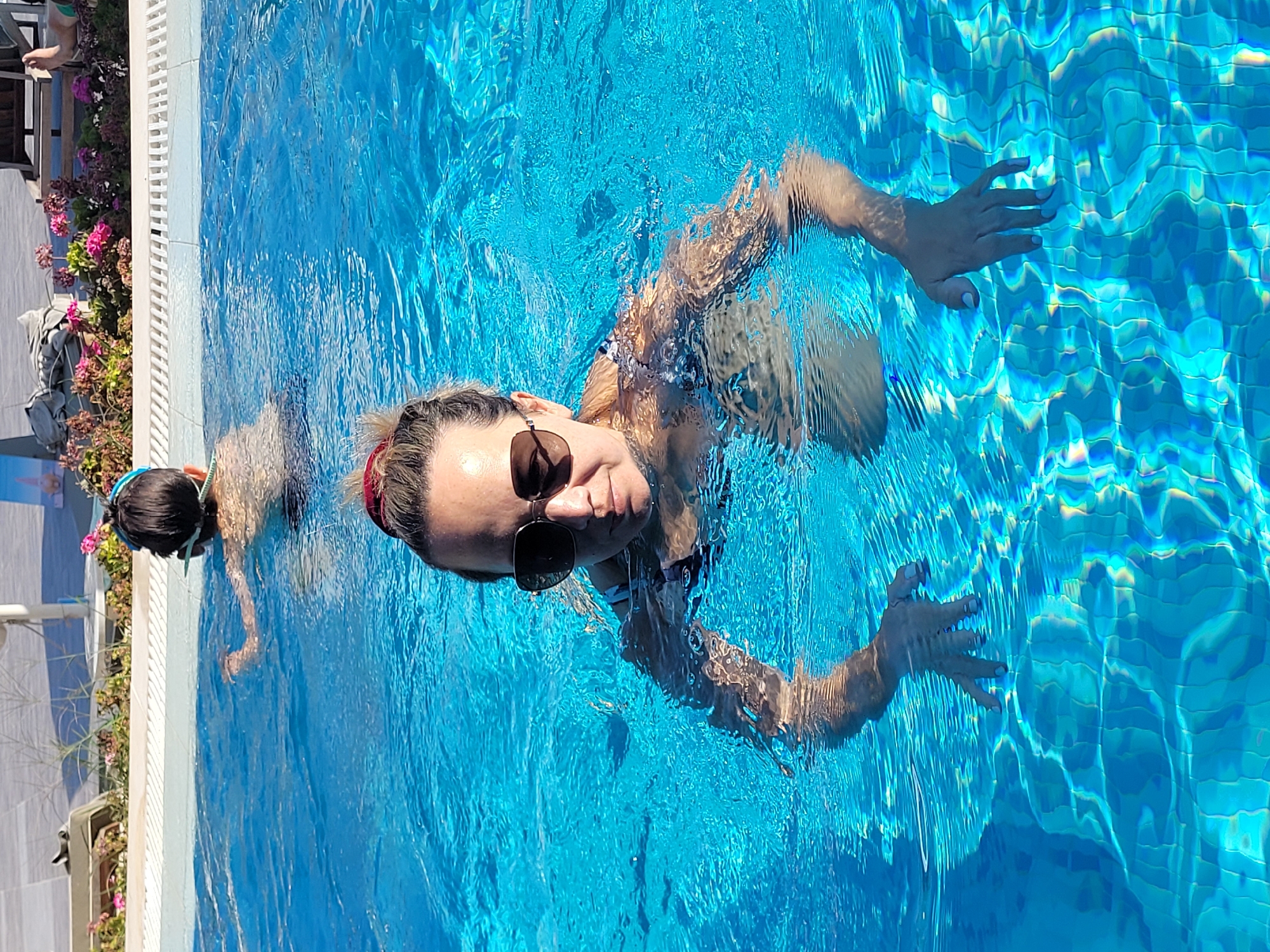 The image shows a woman swimming in a pool. She is wearing sunglasses and her hair is tied up. There is another person in the background, also swimming. The pool is surrounded by a tiled edge and some greenery and flowers. The water is a bright blue and reflects the sunlight. The scene appears to be at a resort or hotel, possibly in Manavgat, Turkey, a popular tourist destination known for its beaches and rivers.