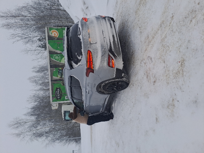A gray car is parked on a snowy road. Snow covers parts of the car, especially the rear. A person is visible near the driver's side door, possibly brushing snow off the car. A large green truck with lettering in Cyrillic is parked behind the car, near bare trees. The background sky is overcast.