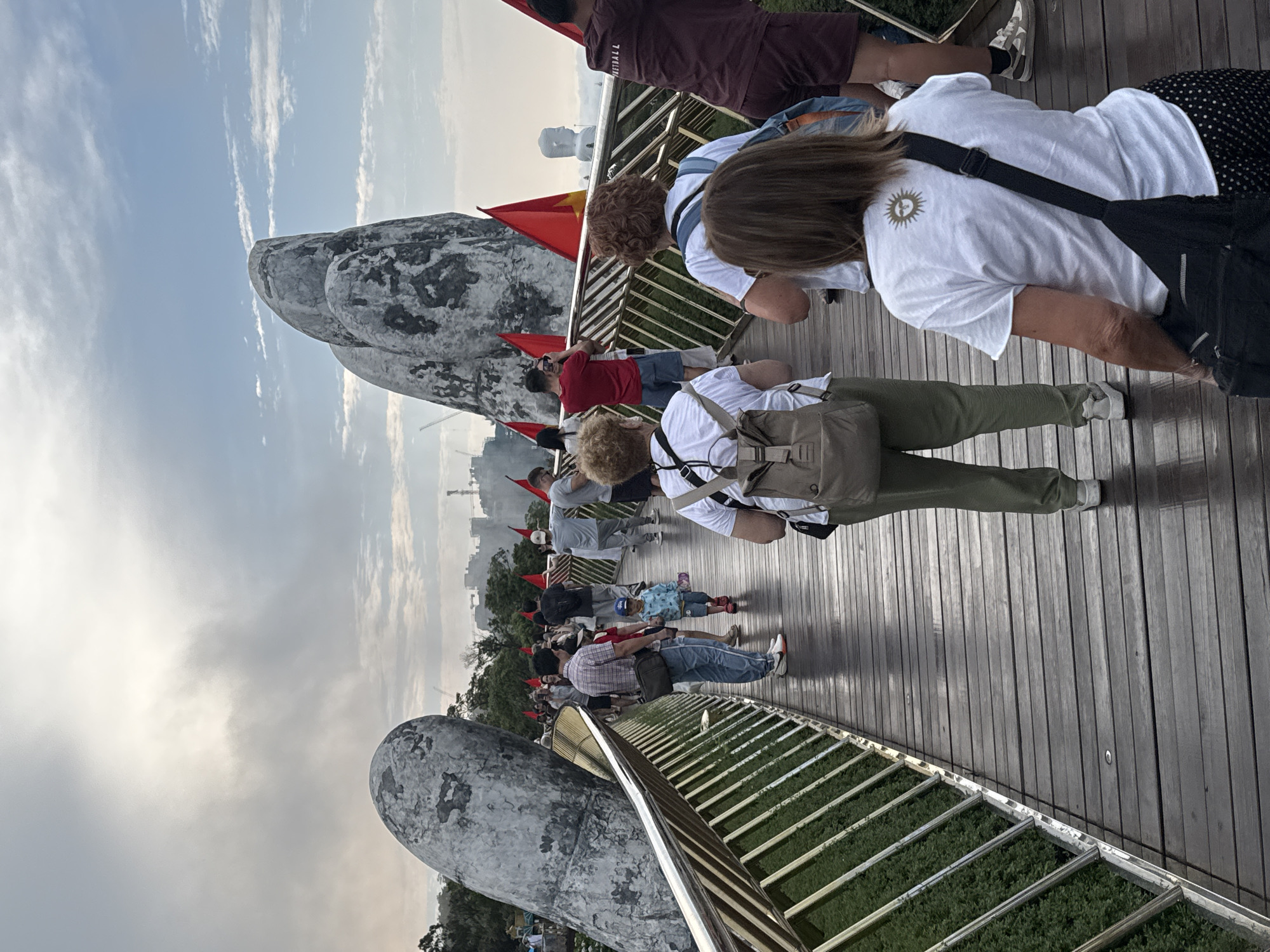 An elevated, crowded pedestrian walkway, commonly known as the Golden Bridge (Cầu Vàng) in Ba Na Hills, Đà Nẵng, Vietnam, is the central focus of the image. The bridge is uniquely supported by two colossal, weathered gray stone hands that appear to emerge from the hillside, holding up the wooden-planked path. Metal railings line the edge of the bridge, with dense green foliage visible below.
Numerous people, a mix of adults and at least one child, are seen walking along the bridge in casual attire. They appear to be tourists, with many carrying backpacks or bags. Several individuals are observing their surroundings, and one person in a red shirt is holding a mobile phone up, likely taking a photograph. Red Vietnamese national flags with yellow stars are intermittently placed along the length of the bridge.
In the background, a large, white Buddha statue can be discerned, a prominent feature of the Linh Ung Pagoda at Ba Na Hills. The sky is partly cloudy, with streaks of white clouds against a lighter blue, suggesting a daytime scene, possibly late afternoon given the soft lighting. The overall mood is one of active sightseeing. Visible text includes "KETBALL" on a person's maroon shorts and a distinct stylized sun symbol on the back of a white t-shirt worn by a person in the foreground.
