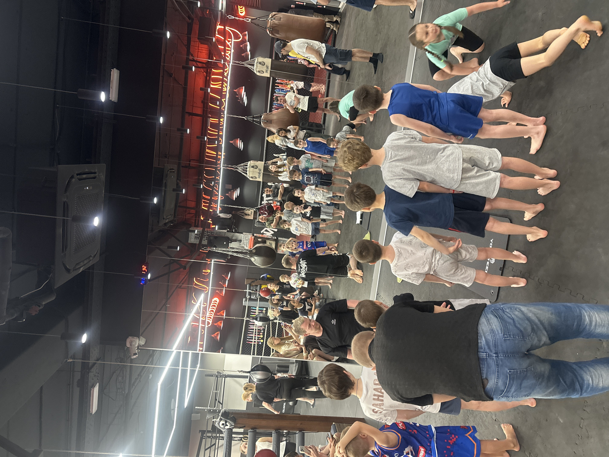 A group of children and adults are arranged in rows on a black mat in what appears to be a gym or fitness center, likely in Perm, Russia. Several punching bags hang from the ceiling, and red neon signage is visible on the wall. The people are barefoot, and some are wearing sportswear.