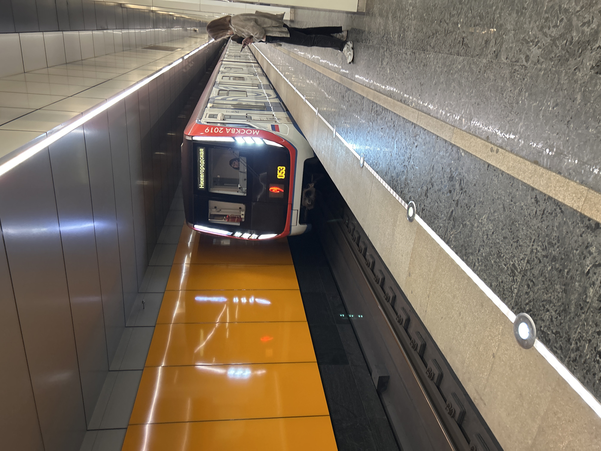 A train with "Нижегородская" and "МОСКВА 2019" written on it is stopped at a subway station. The train is red and white. There is a woman standing on the platform, looking at her phone. The platform is made of stone and has a white line indicating the edge. The station has orange walls and a metallic ceiling with lights.
