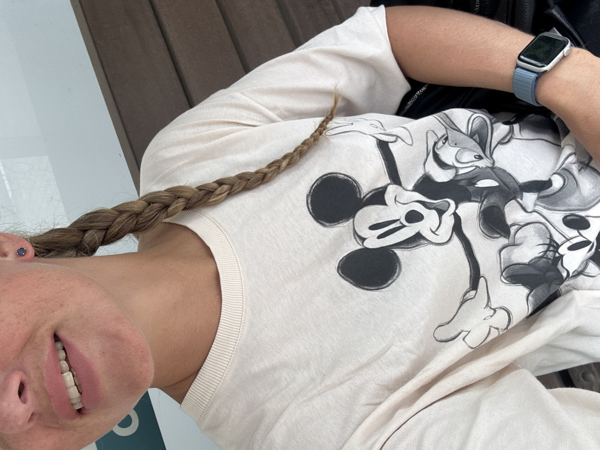 A close-up, medium shot captures a woman from the shoulders up, sitting outdoors. She is wearing a cream-colored t-shirt featuring a black and white graphic of Mickey Mouse and Donald Duck. Her right arm, adorned with a blue smartwatch, rests on her lap. A braid of her blonde hair falls over her left shoulder. The background shows a blurred wooden surface and a white paneled wall. The scene is set in Razdolnoye, Russia.