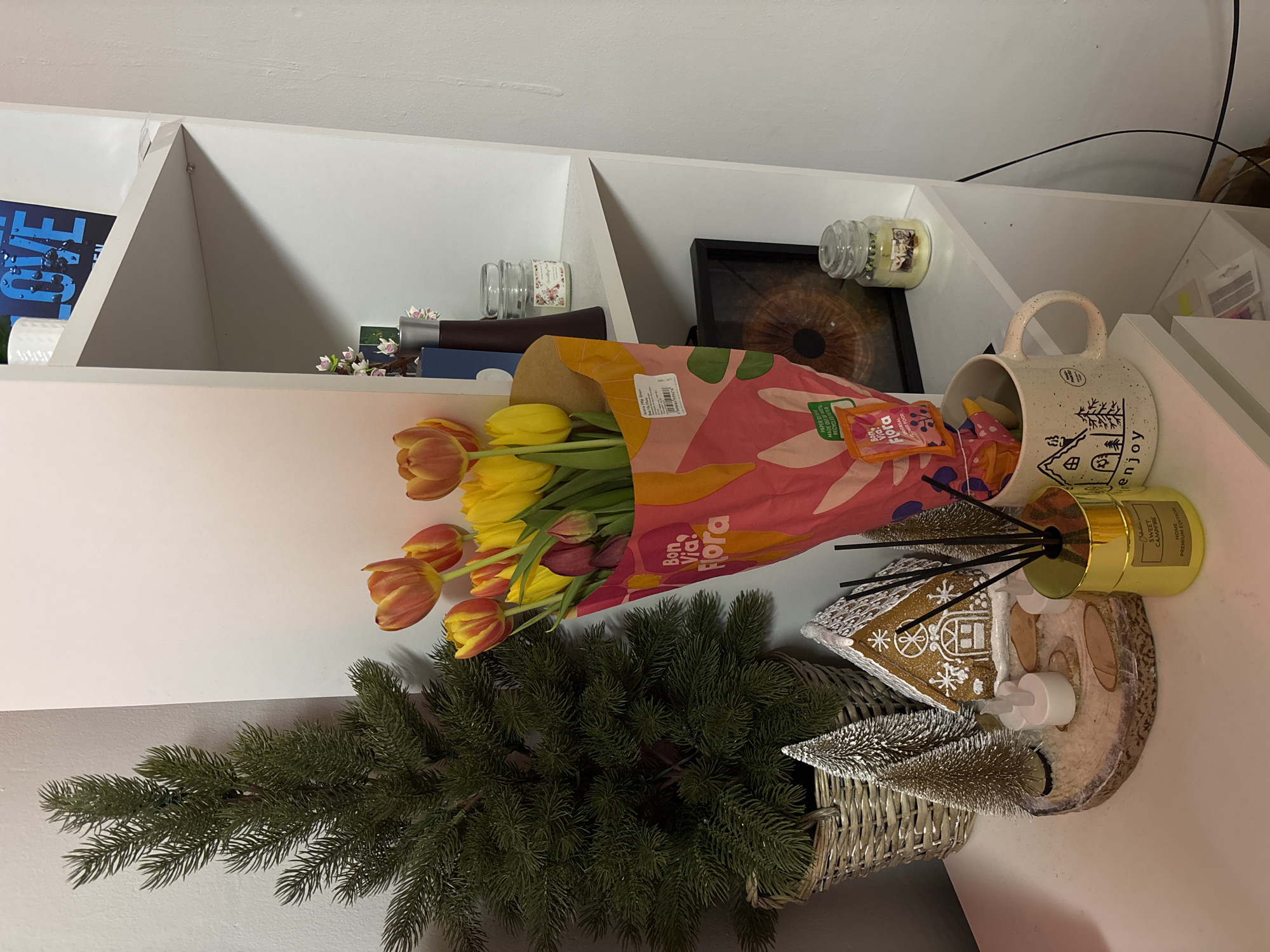 The scene is a close-up still life on a white shelf. Dominating the frame are a bouquet of tulips, predominantly yellow with some orange and purple, wrapped in a pink paper with the brand "Bon Via Flora". Next to the flowers is a decorative evergreen tree in a wicker basket, a miniature silver Christmas tree, a decorative gingerbread house, and a "Sweet Campfire" reed diffuser. A speckled mug with a winter scene and the word "enjoy" is visible. The shelf also holds various objects: books, candles, and a framed eye print. The location and city of Zlatitsa, Bulgaria are not evident from the image.