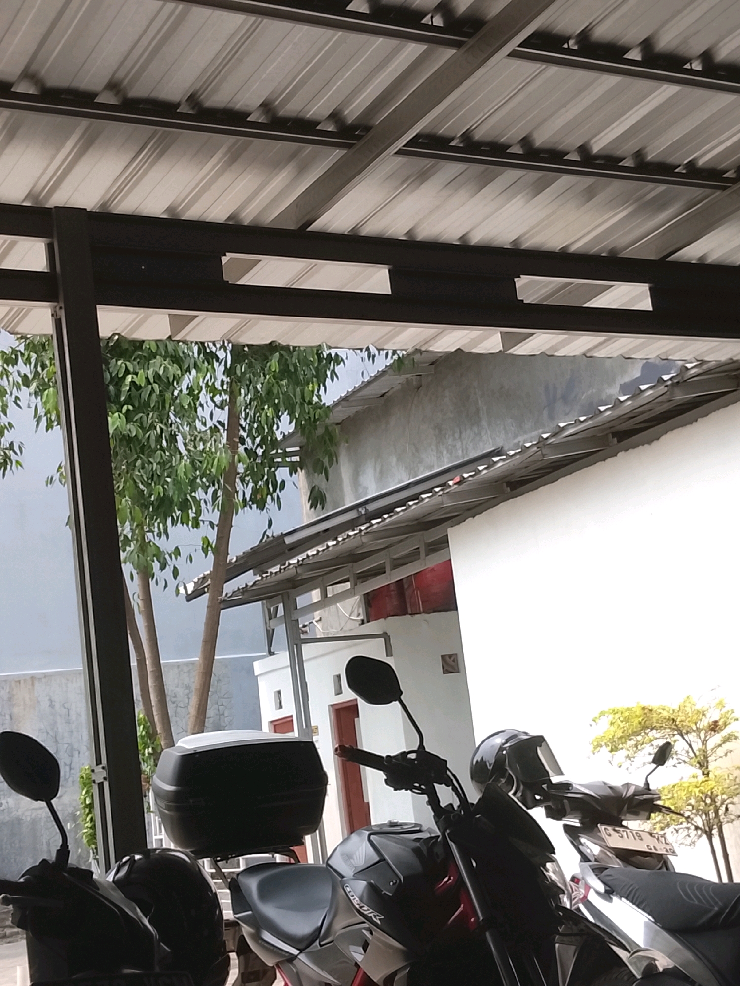 The image depicts a scene outdoors, possibly a parking area under a metal roof structure, located in the city of Slawi, Indonesia. Two motorcycles are visible in the foreground, parked next to each other. The motorcycle on the left is mostly obscured, but its black side mirror is clearly visible. The motorcycle on the right is more prominent, featuring a black seat, silver accents, and a license plate with the characters "G 5719 YZ" and a date that appears to be "04-20". A black helmet rests on the handlebars of this motorcycle. Behind the motorcycles, there is a black storage box mounted on a rack.
In the background, several trees with green foliage are present. A light-colored building with a red section is partially visible to the right of the trees. The overall environment suggests an urban or semi-urban setting. The roof structure overhead is made of corrugated metal panels supported by dark-colored beams. The ground is not clearly visible. There are no people actively present in the scene.