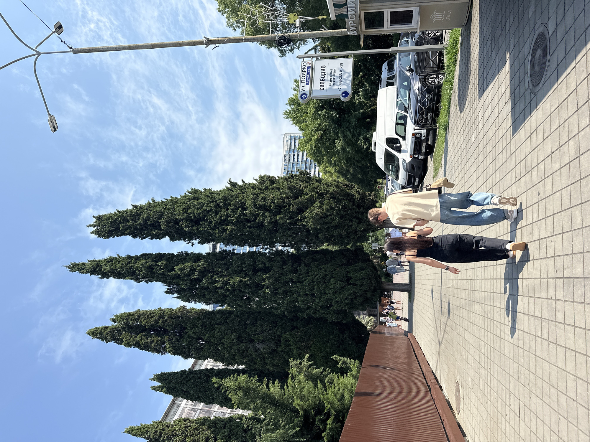 This is a full-shot, eye-level, outdoor, daytime photograph taken on a clear day with some scattered clouds in the sky. The scene depicts a paved pedestrian walkway lined with tall, dark green cypress trees on the left and a row of parked vehicles on the right.
In the foreground, two individuals, a male and a female, are walking away from the camera and towards the left side of the frame. The female, on the left, is wearing a black sleeveless top and black pants. The male, on the right, is wearing a light beige short-sleeved shirt and light blue jeans. He is carrying a tan bag over his right shoulder.
The walkway is constructed of square concrete tiles. To the left of the walkway, a brown metal fence runs along the edge of the frame. Behind the fence, more green trees are visible.
On the right side of the walkway, a row of parked vehicles is visible, including a white cargo van, a white SUV, and a dark gray sedan. In the background, there are multi-story buildings and more trees.
A lamppost with multiple lights stands on the right side of the walkway. Attached to the lamppost is a street sign indicating "ул. Поярко Poyarko str." below which is a sign that reads "СВОБОДНО" (Free) with a phone number "+7-988-233-81-18". Further down the walkway, a small kiosk or booth is visible with the word "СОЧИ" written vertically on its side.
Other pedestrians are seen walking in the background on the walkway, some further away and smaller in scale. The overall environment suggests a public area in Sochi, Russia, likely a street or promenade.