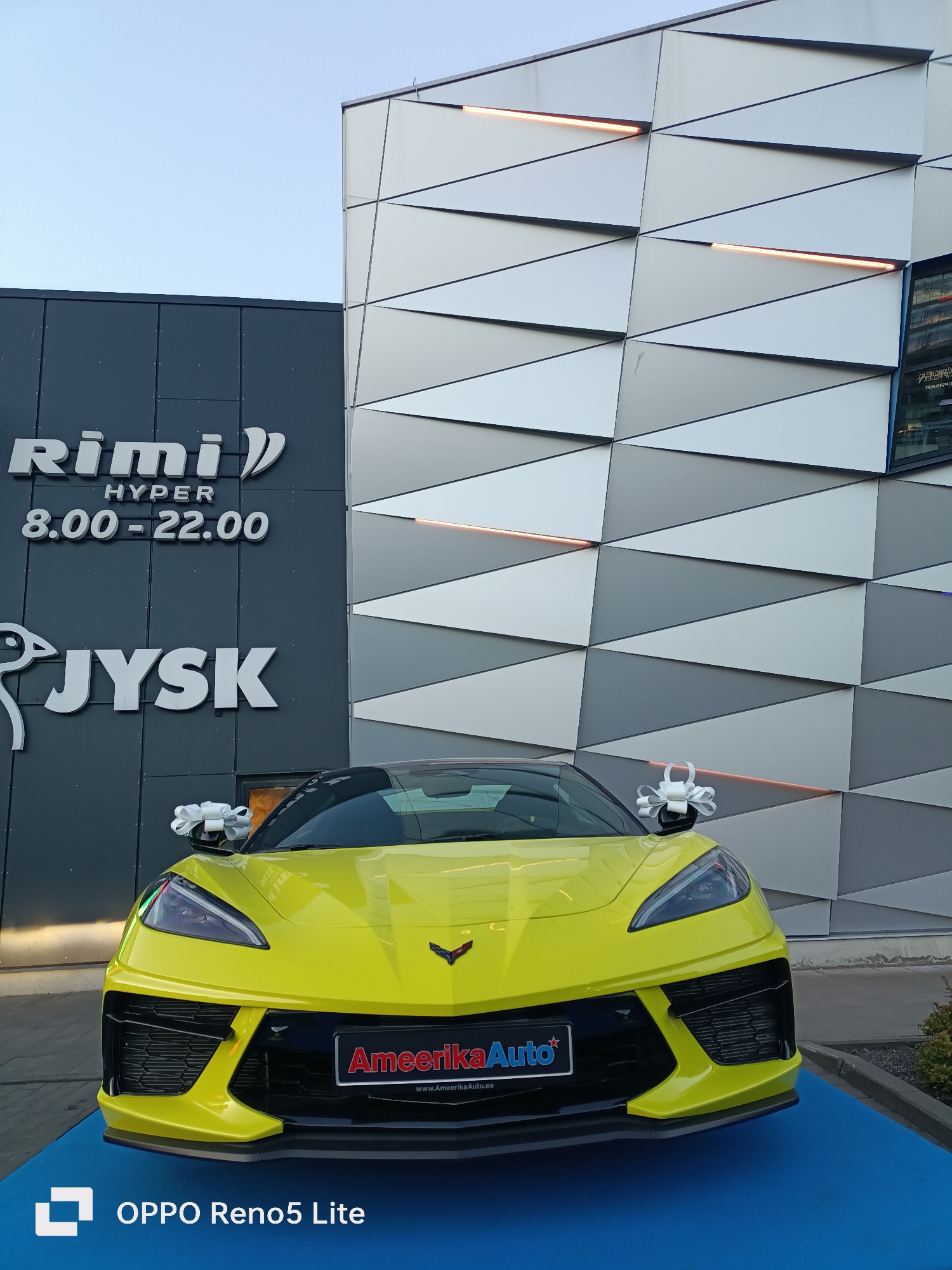 The image shows a bright yellow Chevrolet Corvette parked in front of a modern building with a geometric facade. The building features signs for "Rimi Hyper" and "Jysk". The Corvette has a large white bow on its hood, suggesting it is a gift or a display. The surrounding environment appears to be an urban setting, possibly a shopping center or dealership, with a blue platform beneath the car. The image is a photograph taken with an OPPO Reno5 Lite.