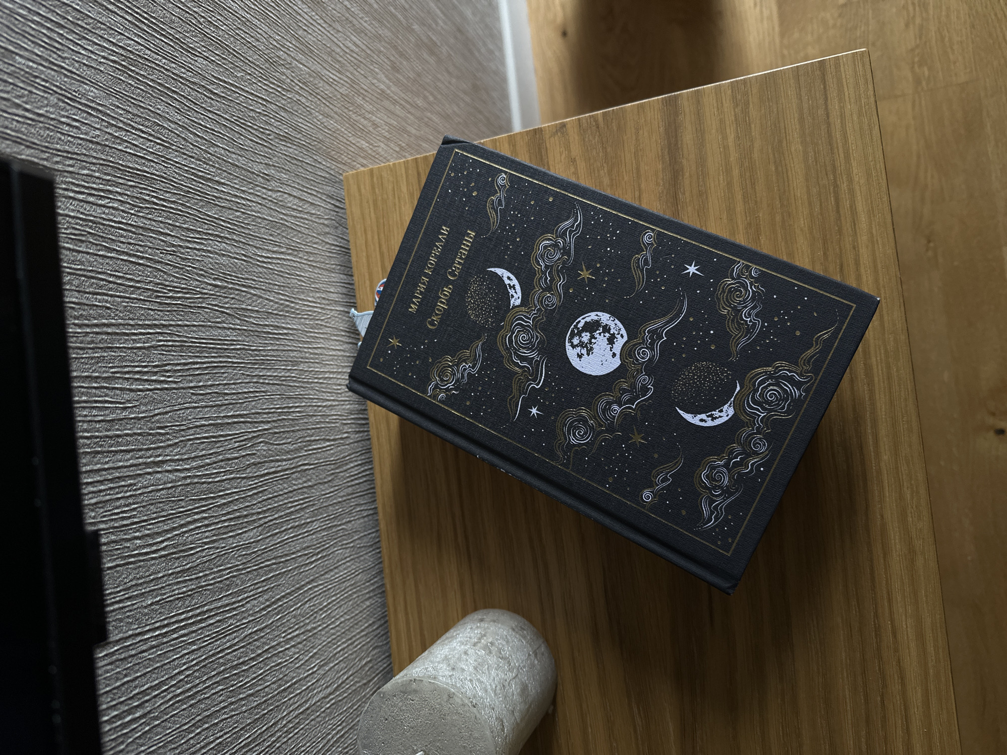 A top-down, high-angle shot depicts a black hardcover book, titled "The Sorrow of Satan" by Mariya Korolev, resting on a light brown, wooden surface, possibly a side table. The book cover is decorated with gold-colored celestial motifs, including moons, clouds, and stars. A small, cylindrical gray object is positioned near the book on the wooden surface. To the left, a gray, textured wall is visible, as well as the corner of a black object, possibly a television. The image does not reveal the location as Yerevan, Armenia.