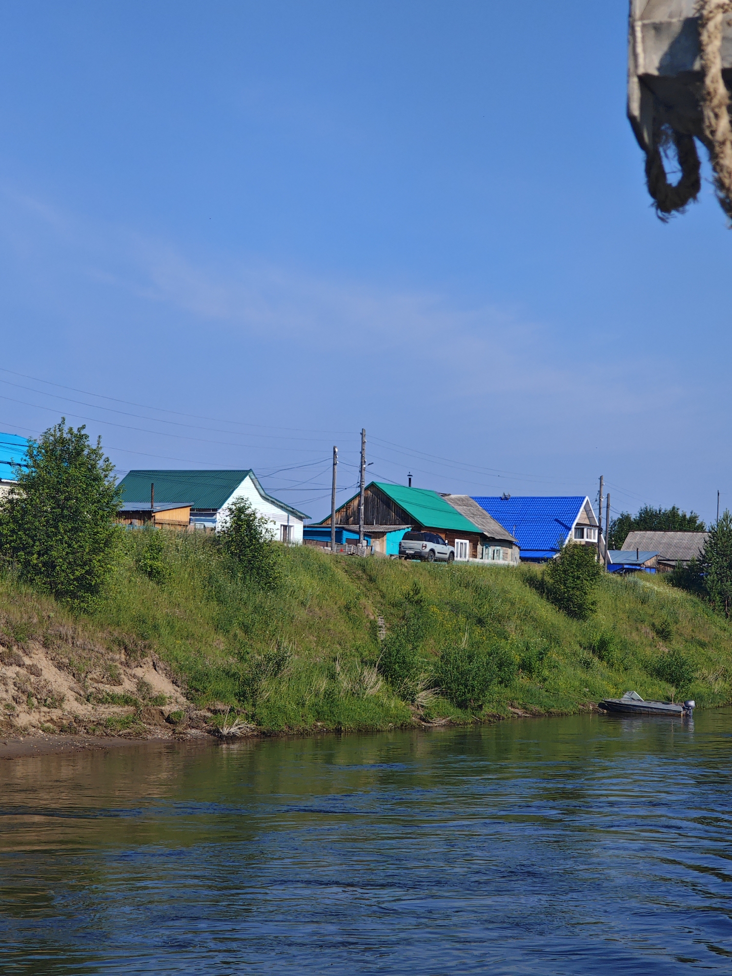 In Galanino, Russia, a serene river flows calmly under a clear blue sky. The riverbank, covered in lush green grass and dotted with trees, slopes upwards towards a row of houses. These houses, characterized by their colorful roofs in shades of green and blue, stand as a charming testament to rural architecture. A silver car is parked in front of one of the houses, suggesting a quiet, residential atmosphere. A small motorboat is tethered to the shore, ready for a leisurely ride on the water. The scene is peaceful and idyllic, evoking a sense of tranquility and the simple beauty of nature.