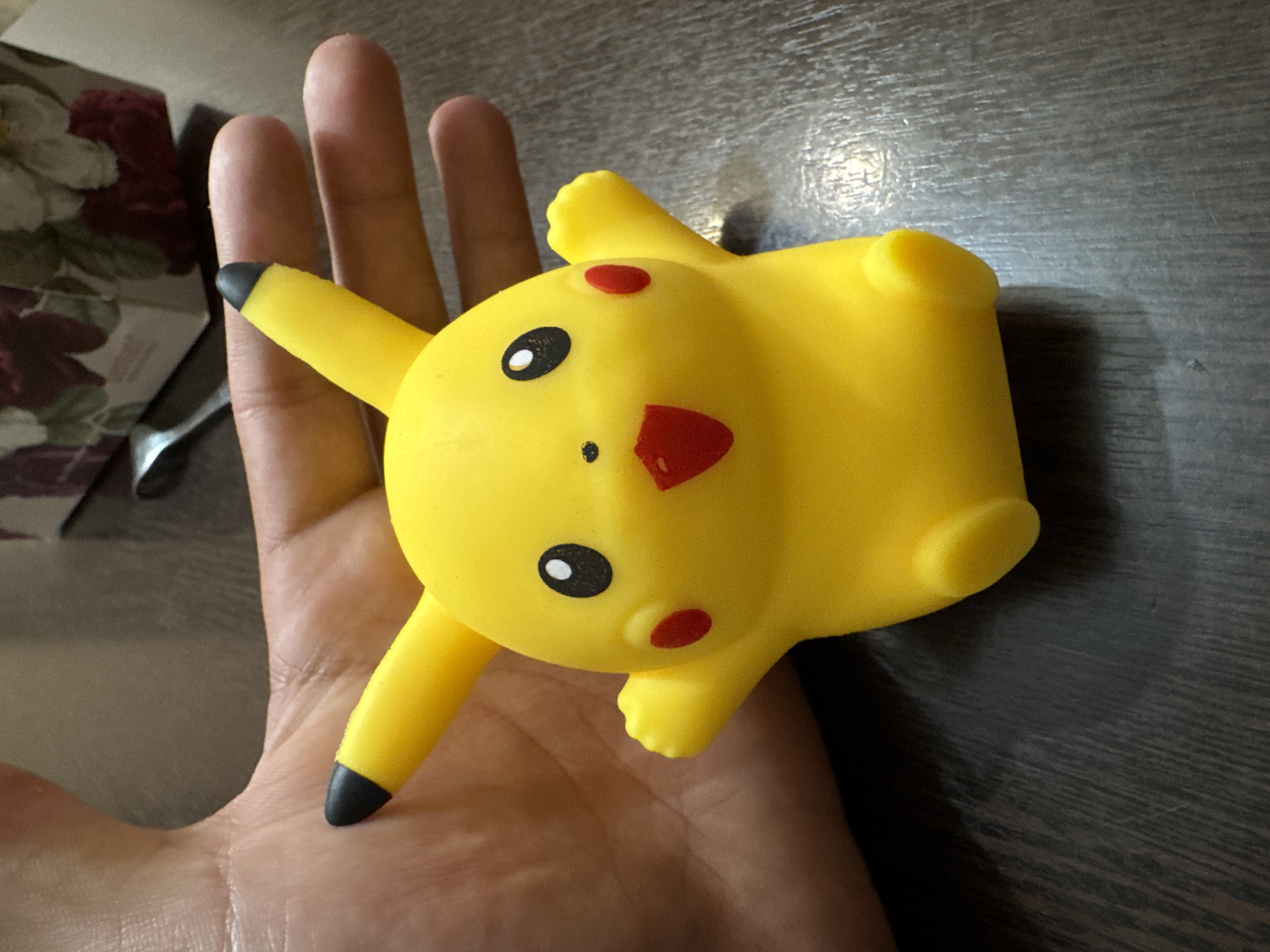 A yellow rubber Pikachu toy rests in the palm of a human hand, its head facing the camera. The toy has black-tipped ears, black eyes with white highlights, red cheek spots, and a red mouth. The background shows a dark, wood-grain surface and a glimpse of a floral patterned object to the left. The scene appears to be indoors, but no location in Yerevan, Armenia, can be determined from the available visual information.