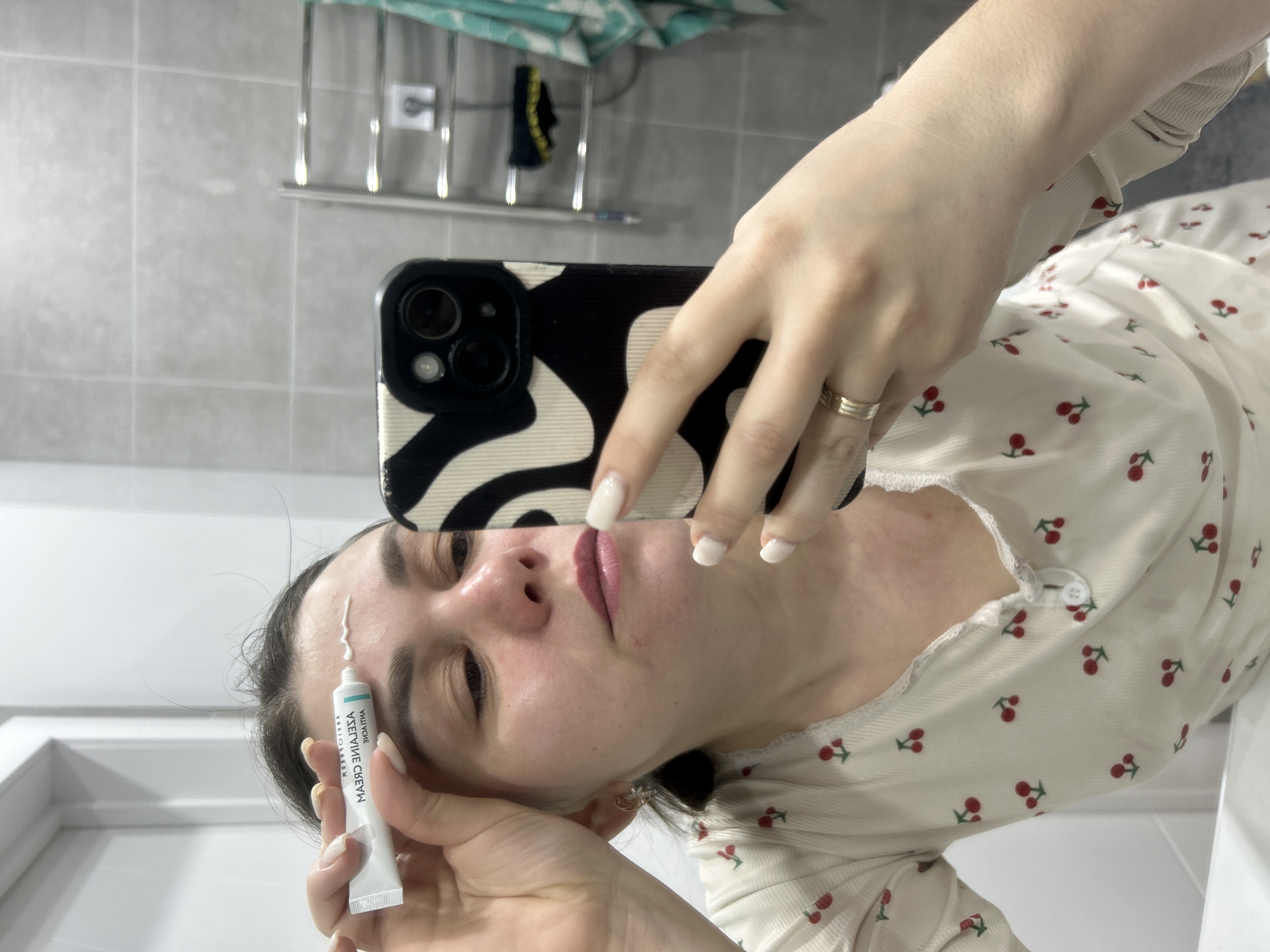 A woman is taking a selfie in a bathroom. She is holding a tube of "AZECLAINE CREAM" and has squeezed some of the cream onto her forehead. The woman is wearing a white top with a cherry print. She has short white nail polish. She is also wearing a wedding band. The background is a bathroom, with tiled walls and a towel rack with items hanging on it. Location cannot be determined.