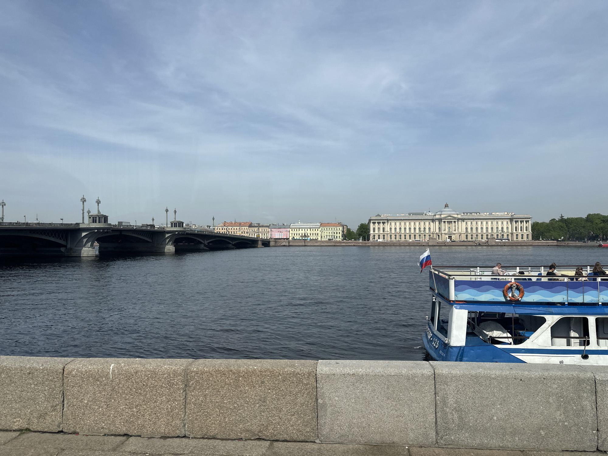 The image shows a waterfront scene in Saint Petersburg, Russia. A gray concrete wall forms the foreground. Behind it is a wide river with a boat in the foreground. The boat is two-tiered with blue and white paint, a Russian flag flying from its stern, and several passengers on the upper deck.
In the background, a bridge spans the river from left to right. Beyond the bridge, a row of colorful buildings lines the waterfront. Further back on the right is a large, pale-colored building with a prominent colonnade. The sky is light blue with scattered clouds.