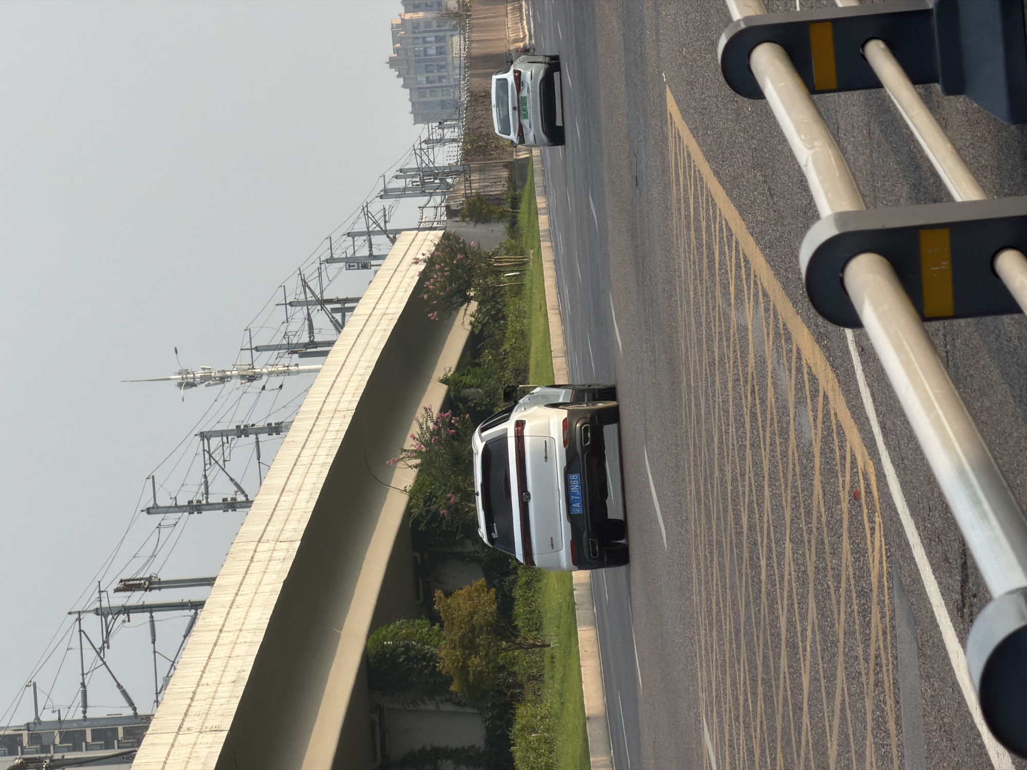 The image captures a street scene in Jiangjin, China, with a focus on vehicular traffic and urban infrastructure. In the foreground, a white SUV with the license plate "A 7JN88" is driving on the right side of the road. Behind it, a white sedan with the license plate "A AF599" is also visible. The road has visible lane markings, including yellow and white lines, and a grid pattern of yellow lines indicating a potential hazard or no-stopping zone.
The environment includes a raised overpass or bridge in the background, supported by concrete pillars. Overhead, numerous power lines and utility poles are present, characteristic of urban electrical and communication infrastructure. Vegetation, including trees and bushes, lines the side of the road, with some flowering plants visible. In the far distance, several multi-story buildings can be seen. The sky is a pale, overcast grey. The overall impression is an active urban roadway under a muted sky.