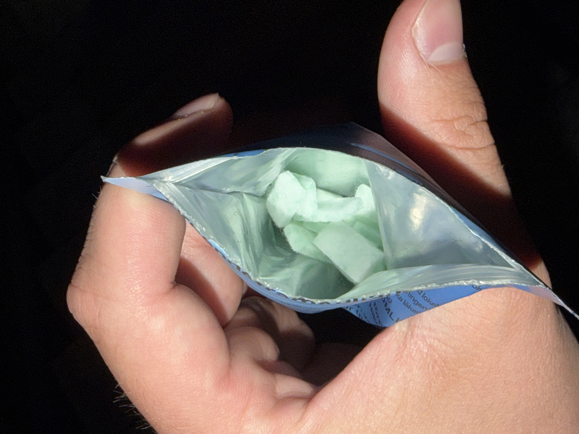 An outdoor close-up shot shows a person's hand holding open a small, light blue, resealable pouch. Inside the pouch are several light green, irregularly shaped pieces of what appear to be candy or gum. The background is completely black. The pouch is partially transparent, revealing the contents within. The hand is well-lit, with visible skin texture and fine hairs on the fingers. The lighting suggests the photo was taken at night or in a dimly lit area.