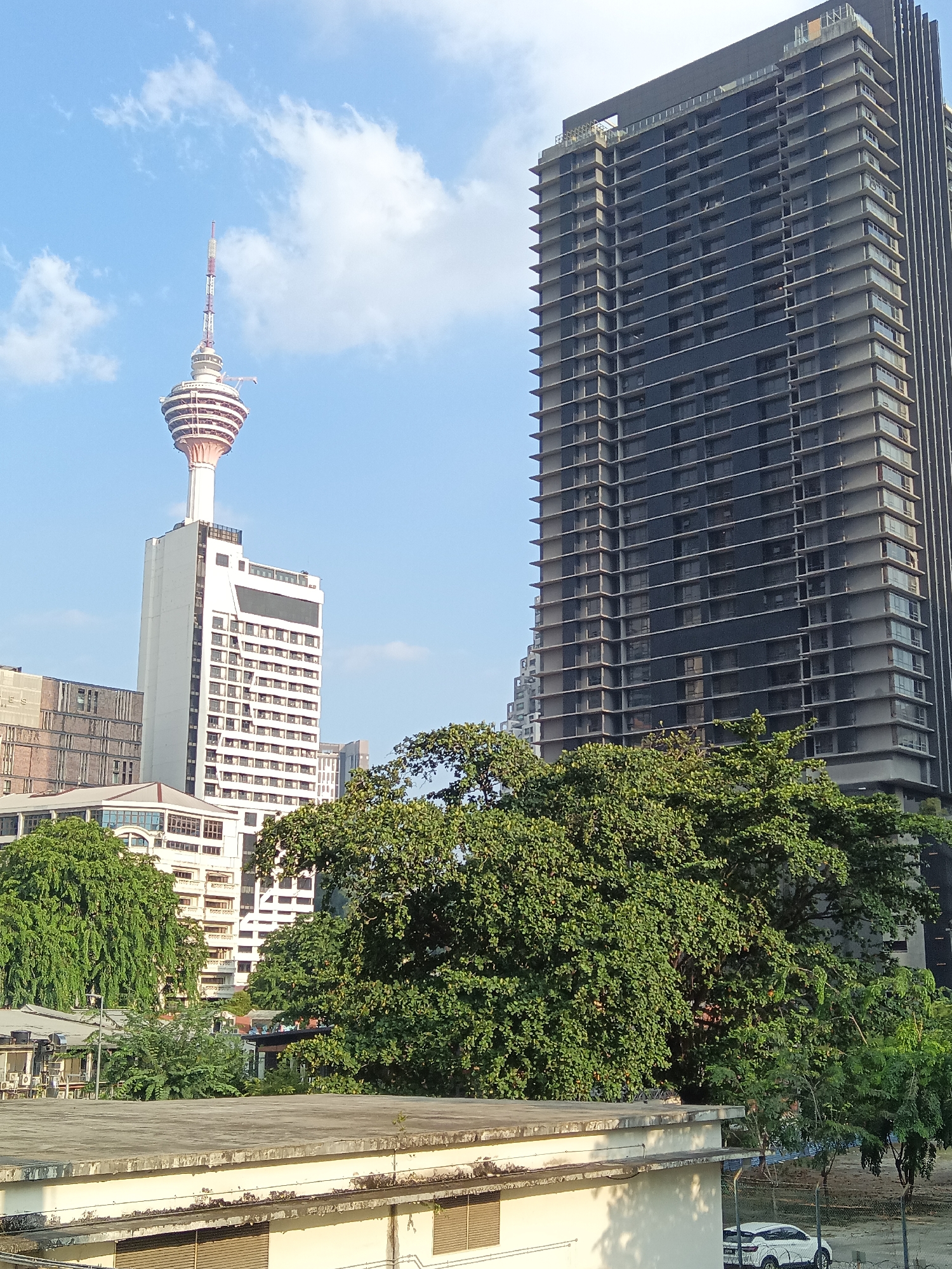 This is a photograph of the Kuala Lumpur Tower and a modern skyscraper in Kuala Lumpur, Malaysia, under a partially cloudy blue sky. The Kuala Lumpur Tower, a prominent landmark, is visible on the left, with its distinctive red and white antenna atop a tiered, rounded structure that sits on a white rectangular building. To the right of the tower is a tall, dark, modern skyscraper, appearing to be under construction or newly built, with a repeating pattern of horizontal balconies and dark glass windows. In the foreground, lush green trees partially obscure the lower portions of the buildings. A flat, concrete rooftop is visible in the immediate foreground. Further back, other buildings and more trees can be seen, suggesting an urban environment. The image captures a slice of the Kuala Lumpur skyline, showcasing a mix of iconic structures and contemporary architecture amidst natural foliage.