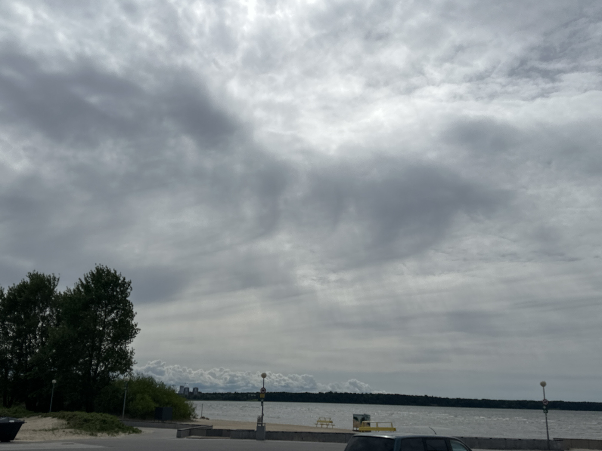 The image shows a cloudy sky over a body of water, likely the Baltic Sea, near Tallinn, Estonia. In the foreground, there is a paved area with a few trees on the left and some urban structures in the distance. The water is choppy and reflects the overcast sky. There are no people visible in the image. The overall scene suggests a slightly overcast day in an urban waterfront area.