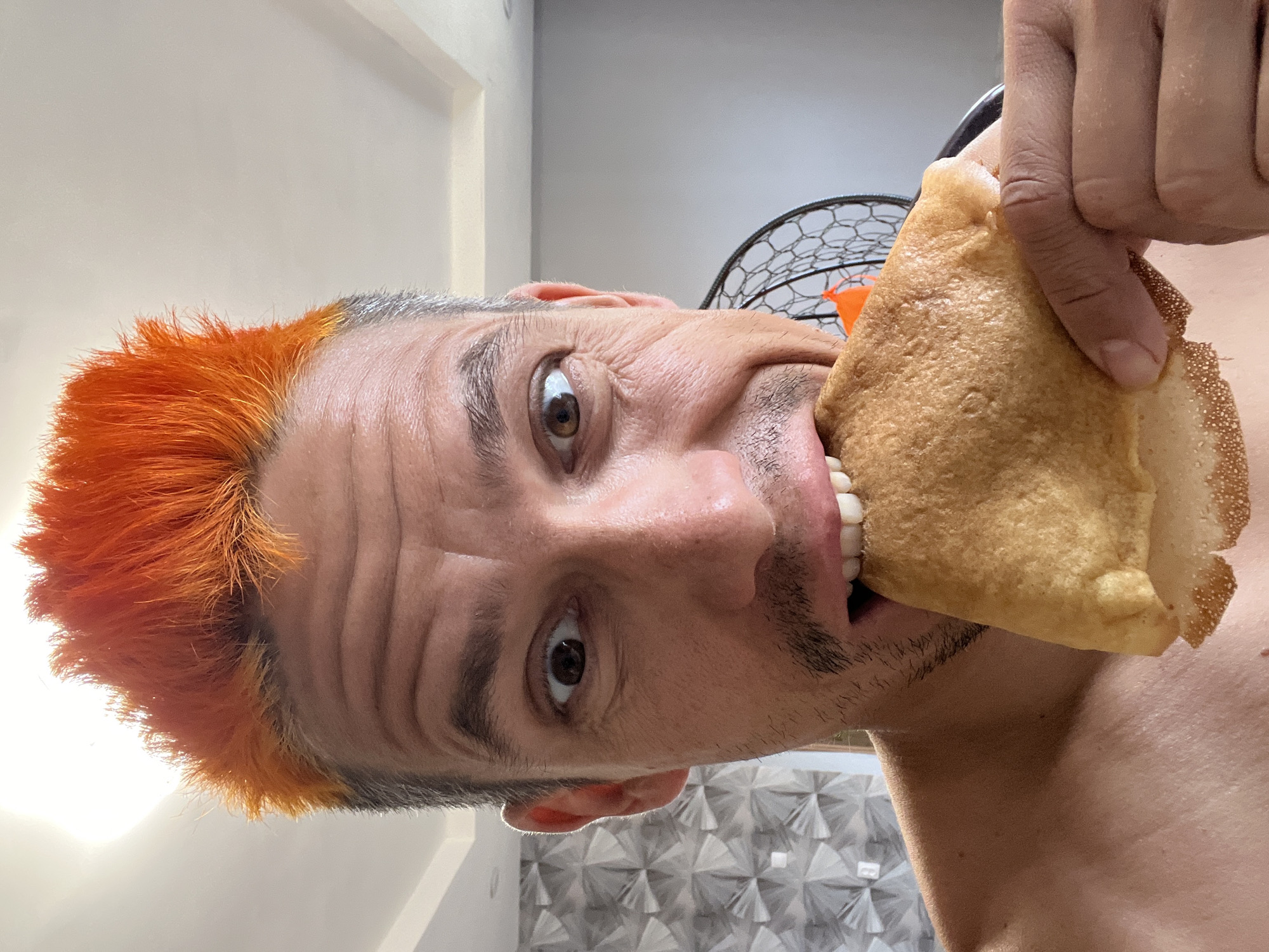 A man with orange spiked hair is biting into a large, flat, golden-brown pastry. He is holding the pastry with his right hand. His eyes are wide. A portion of his bare shoulder is visible. The background includes a white ceiling, a gray wall, and a glimpse of a black wire fan. The location is Chaikovsky, Russia.
