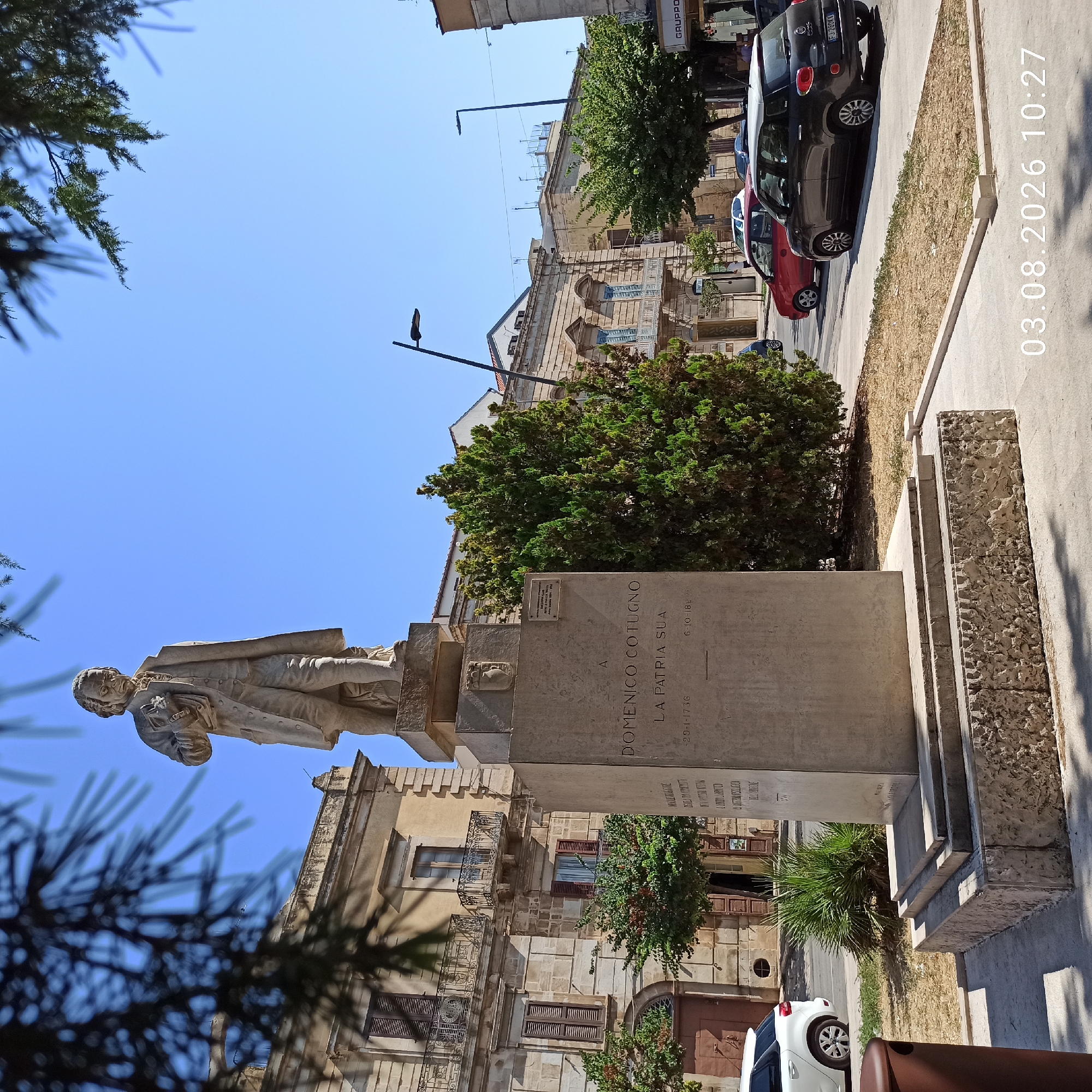 The image depicts a statue of Domenico Cotugno in Ruvo di Puglia, Italy. The statue is made of stone and stands on a pedestal in a public square. In the background, there are several buildings, some of which appear to be old and historic, with classical architectural elements. There are also trees and streetlights visible, suggesting an urban environment. A few cars are parked or driving on a street to the right of the statue, and there are also some pedestrians in the distance. The sky is clear and blue, indicating a sunny day. A timestamp overlay in the lower right corner reads "03.08.2026 10:27". The overall scene is of a well-maintained public space, likely a town square or plaza, commemorating a historical figure.