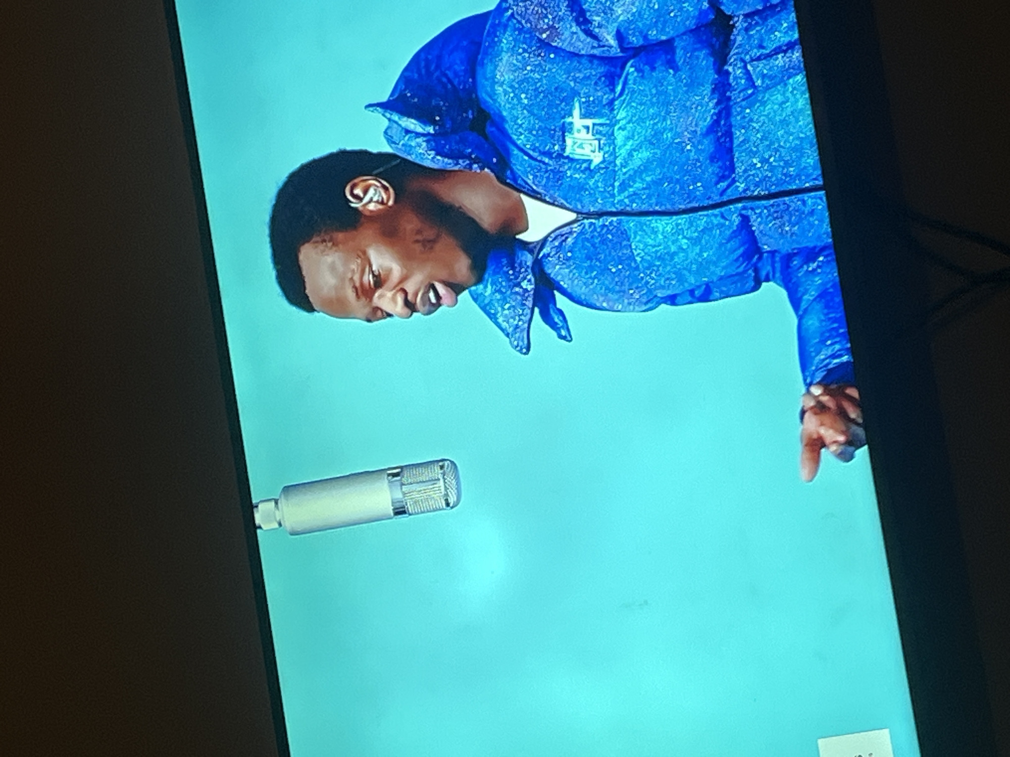 The image shows a close-up of a man wearing a blue, puffy jacket adorned with glitter. He has dark hair and is looking to his left with his mouth slightly open, as if speaking or singing. A microphone is visible above and to the left of his head. The background is a solid, bright blue color. The man's left hand is extended towards the bottom of the frame, with his index finger pointing. A small white object with text is partially visible in the bottom left corner. The scene appears to be on a screen, possibly a television or monitor, indicated by the dark bezel at the top and bottom. There is no indication of the city of Gura Top, Nigeria, in the image or its content.