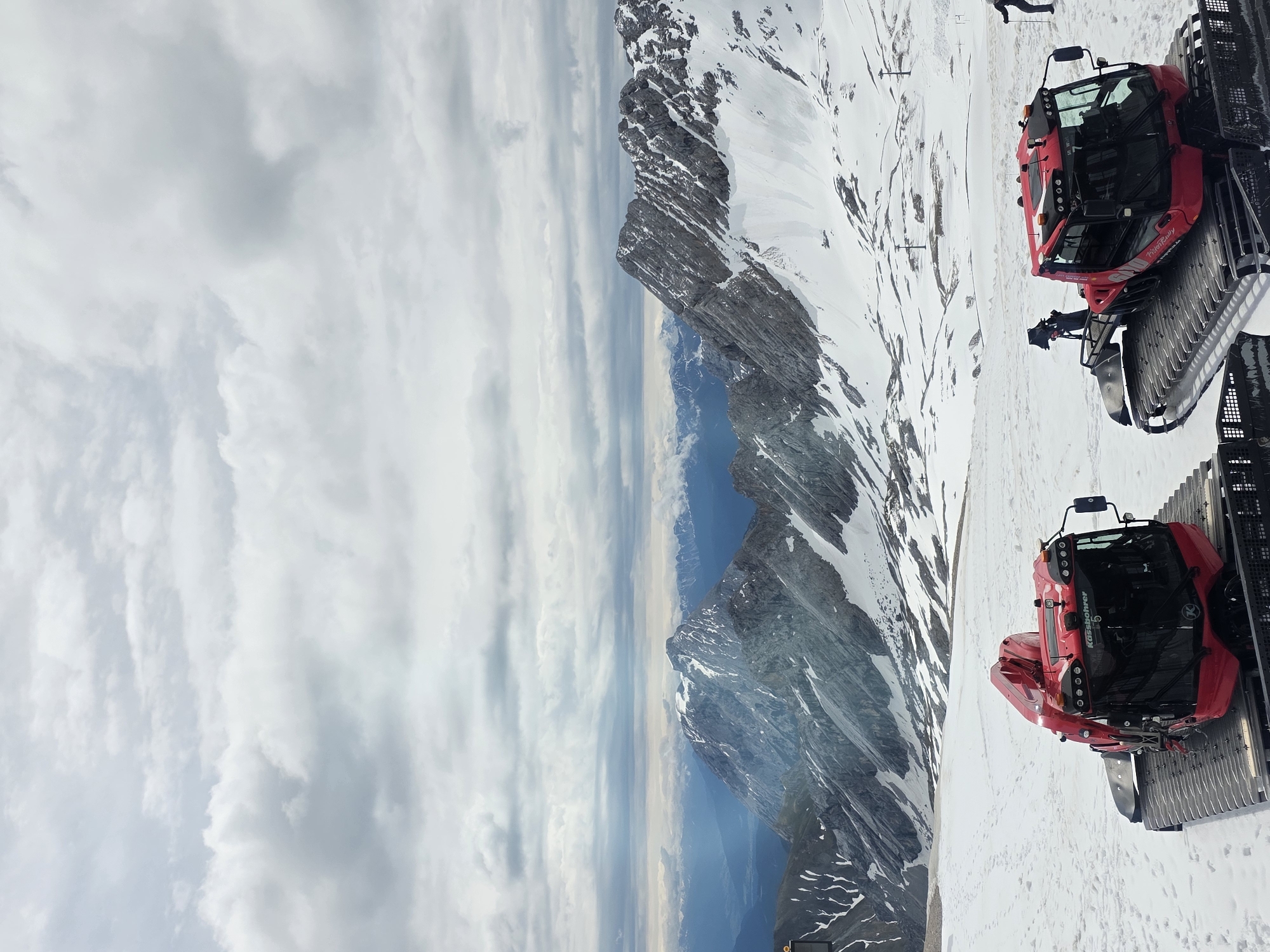 A wide panoramic view captures a snow-covered mountain landscape under an overcast sky, observed from an elevated position. The scene is located at Zugspitze Glacier, Bavarian Alps, Germany. In the immediate foreground, two red PistenBully snow groomers are parked on a vast expanse of snow. The groomer on the left, displaying "Kässbohrer" and the number "5" on its front window, faces slightly right. The groomer on the right, positioned further back and to the right, features "PistenBully" and "ZUGSPITZ GLETSCHER" branding on its side. Both machines are equipped with snow blades and wide tracks. To the far right edge of the frame, a person wearing dark attire and holding poles stands in the snow. The snowfield in the foreground shows numerous footprints. Beyond the groomers, the terrain slopes upwards, revealing rugged, grey rock formations partially covered with snow. A prominent snow-capped peak rises in the mid-ground, surrounded by lower, jagged ridges. Distant mountain ranges, appearing blueish and less defined, stretch across the horizon. The sky is heavily overcast with thick, grey clouds, yet a faint beam of light penetrates the clouds in the upper-left mid-ground, subtly illuminating a distant valley. Ski lift infrastructure, including poles, is sparsely visible within the mid-ground snowfield.