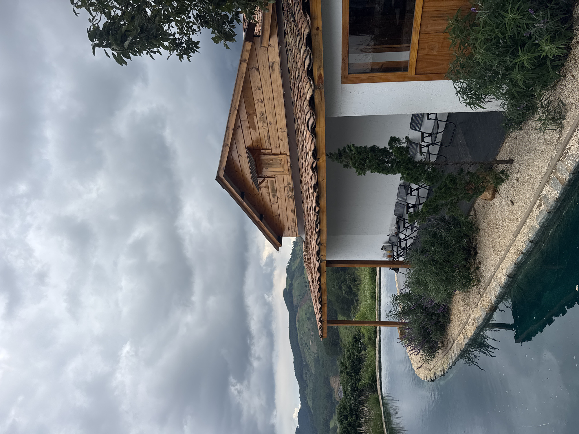 A side-view, medium shot captures a picturesque scene in Thôn 3, Vietnam, with a focus on a charming wooden-accented building overlooking a serene, curved swimming pool. The building features a distinctive terracotta tile roof and white walls, with a wooden eaves and a small, louvered window on the upper level. To the right of the building, a wooden door with a glass pane is visible, reflecting the cloudy sky. Several dark folding chairs are arranged under the building's covered porch, suggesting a relaxing seating area.
The swimming pool, with its dark blue water, gracefully curves along the foreground, bordered by a textured stone edge and a bed of light-colored gravel. Lush green and purple flowering plants line the edge of the pool, adding a touch of natural beauty. In the background, rolling green hills, covered in dense vegetation, stretch towards a dramatically cloudy sky. The overall atmosphere is tranquil and invites relaxation amidst stunning natural scenery.
