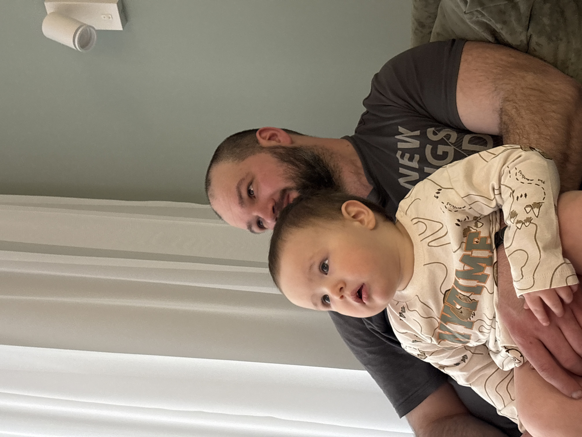 A man with a beard holds a baby wearing a patterned pajama top. They are positioned in front of a window with blinds. A light fixture is visible on the ceiling. The image does not contain sufficient information to determine the location as Izhevsk, Russia.