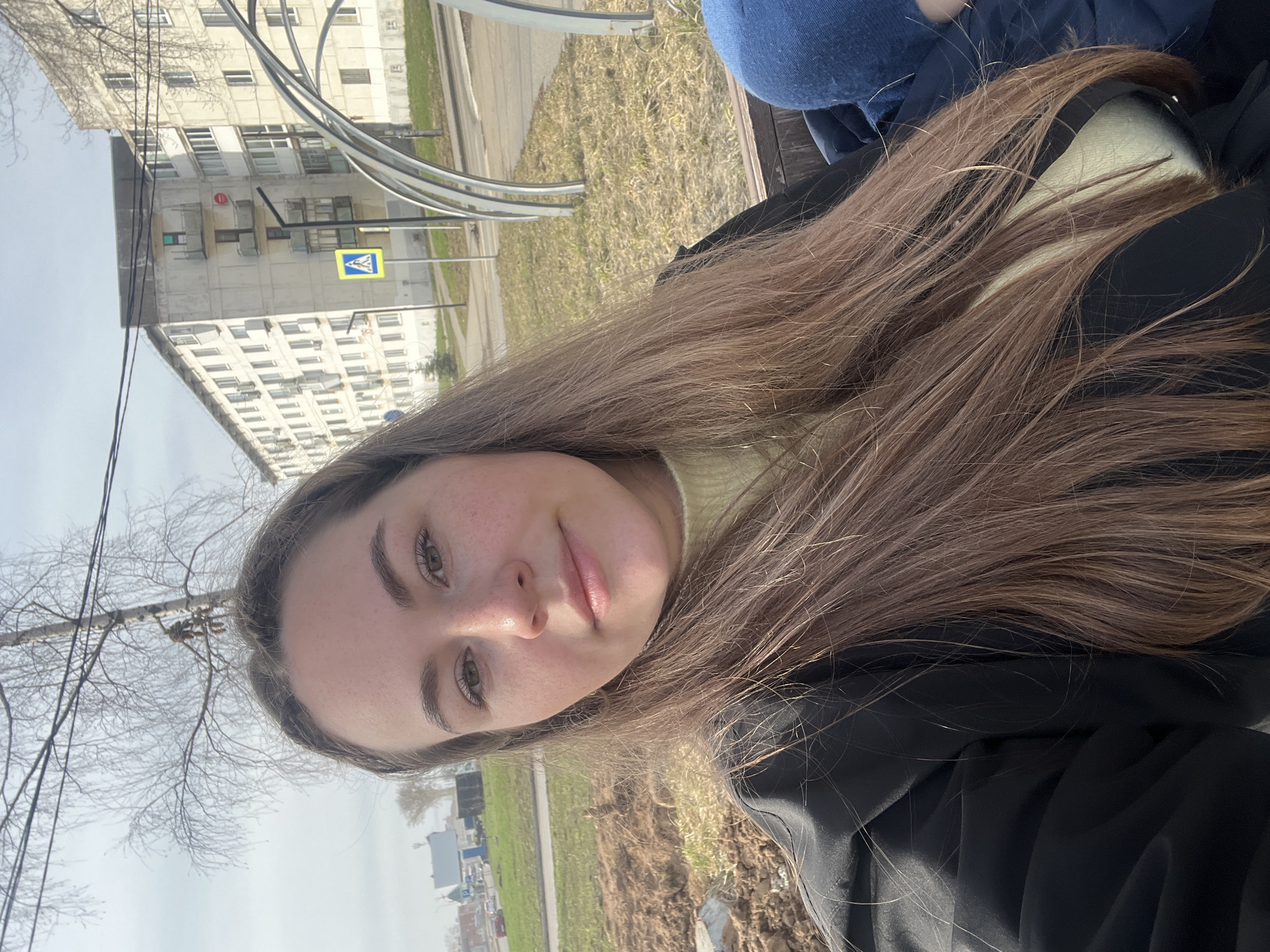 The photo shows a woman with long brown hair, wearing a black jacket and a yellow top. She is outdoors, likely sitting on a bench in a city park. The background includes a residential building with multiple windows, a yellow pedestrian crossing sign, a grassy area with bare patches, and bare tree branches. The photo was taken during the day with natural light. Based on these details, the photo was likely taken in Chaikovsky, Russia, given the provided context.