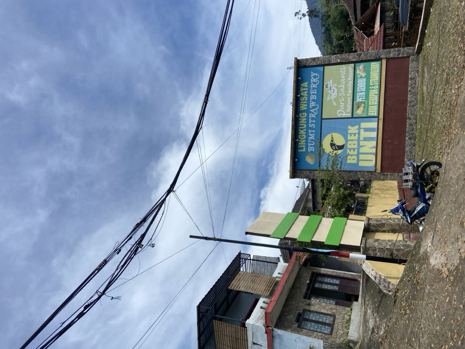The scene is a street corner in Rancabali, Indonesia, located at latitude -7.12 and longitude 107.432. The image shows a partially obscured blue and white sign, with text in Indonesian, on a stone wall. The text on the sign reads “Lingkung Wisata Bumi Strawberry” (Nature Tourism Strawberry Land). The sign is directly behind a black motorcycle parked on a steep, dirt slope, and beside a green and white sign. The motorcycle is facing away from the camera. In the foreground, there are leaves scattered on the dirt slope. A small, two-story building, with wood and stone exterior, is on the left side of the image. There are many black electrical wires running overhead. In the background, is a densely forested hillside with green trees. A partial view of a roof of a second building is in the background on the right side of the image. The sky is blue, with scattered white clouds.
