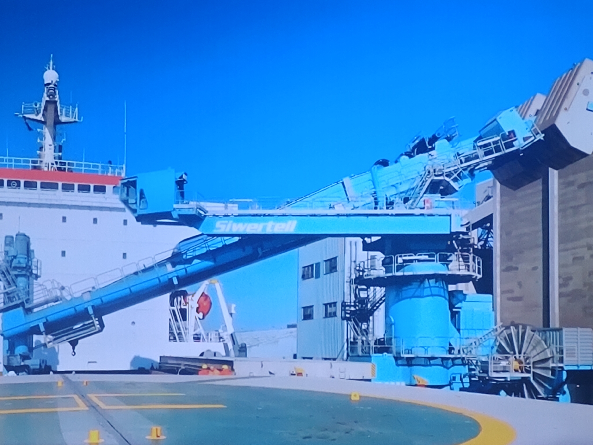 The image shows a large, blue conveyor system, labeled "Siwertell," mounted on a ship. The ship is docked next to a light-colored building with multiple windows, and its superstructure with a mast is visible. The conveyor extends over the ship's deck and appears to be designed for bulk material handling. The scene is outdoors under a clear blue sky. There are no people clearly visible in the image. The ground in the foreground is a green surface with yellow markings, likely a port or industrial area. This scene is consistent with activity at the Port of Brussels, which is located in the municipality of Molenbeek-Saint-Jean - Sint-Jans-Molenbeek, Belgium.