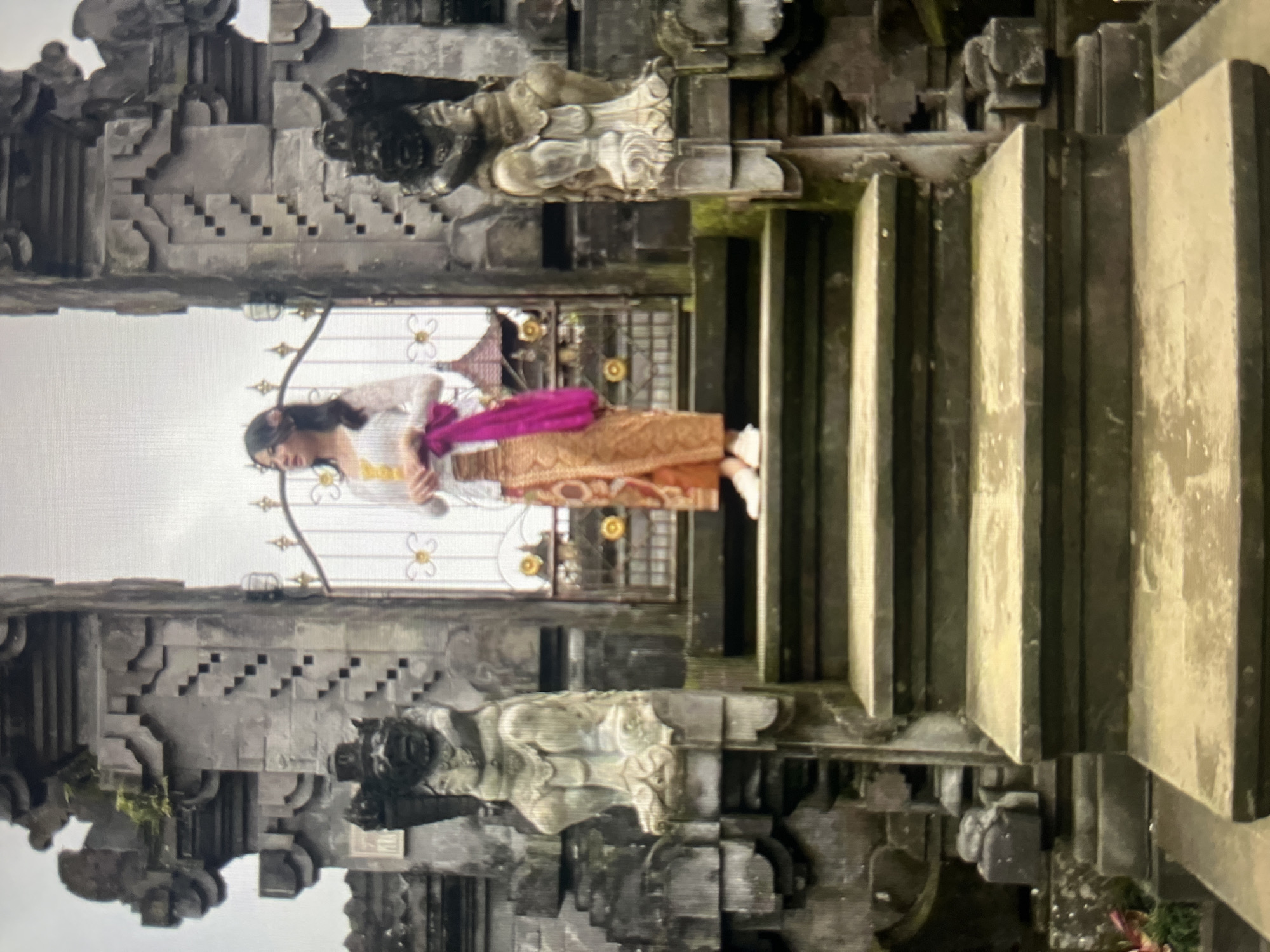 A young woman stands on a stone staircase in front of a gateway. She is wearing a traditional Balinese outfit, including a white lace top, an orange patterned sarong, and a pink sash draped over her shoulder. The gateway is made of stone and features two carved statues on either side, resembling mythical guardians. Behind the woman, a decorative metal gate is visible. The overall setting appears to be a temple or historical site, with weathered stone architecture and a cloudy sky. The image does not contain information about the city of Cirahayu, Indonesia.