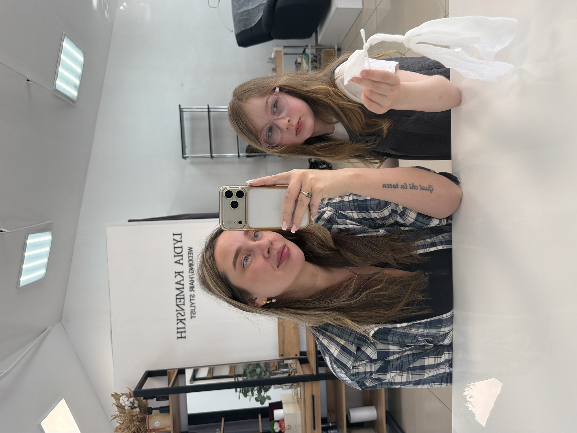 An indoor mirror selfie captures an adult woman and a young girl in what appears to be a hair salon or styling studio in Chaikovsky, Russia. The image is initially rotated 90 degrees counter-clockwise.
The adult woman, positioned on the right (when viewed upright), has long, light brown hair with blonde highlights. She is wearing a blue, green, and white plaid shirt and a ring on her left ring finger. She holds a smartphone (an iPhone with multiple cameras) to take the mirror selfie, looking at her reflection with a slight smile. A tattoo with foreign text, partially visible as "qual chi la tocca" and "socos" (reversed), is on her right forearm.
Beside her, to the left in the reflection, stands a young girl with long, straight, light brown hair. She wears pink-framed glasses and a dark top over a white shirt. The girl holds crumpled white tissues or paper in her right hand and looks towards the camera with a neutral to slightly serious expression.
The background features a clean, modern interior with white walls and ceiling. A prominent sign behind the woman, reflected backward, reads "LYDIA KAMENSKAYA WEDDING HAIR STYLIST," indicating the professional setting. Other visible elements include a black chair, a shelving unit with items like dried flowers and rolls of paper, and rectangular fluorescent light fixtures on the ceiling. A white table or counter occupies the foreground where the subjects are positioned. The scene is well-lit by artificial lights, suggesting an indoor daytime setting.