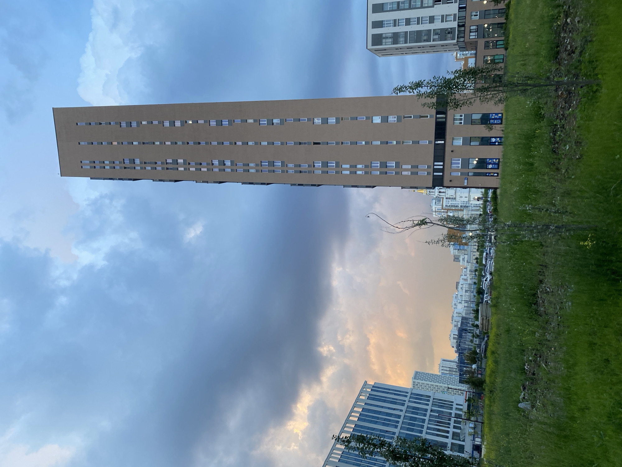 The image captures an urban landscape in Yekaterinburg, Russia, under a dramatic, cloudy sky. In the foreground, a grassy field with some scattered debris leads to a row of modern residential buildings. The most prominent structure is a tall, slender, tan-colored apartment building with numerous small, vertically arranged windows. To its left, a white and blue building with a distinctive facade stands. Further in the background, a cluster of white buildings forms a cityscape, with a slender white spire visible to the left of the tan apartment building. A street with parked cars is visible between the grassy area and the buildings. The overall impression is one of a developing urban area with a mix of contemporary architecture and open green spaces, under a moody, overcast sky.