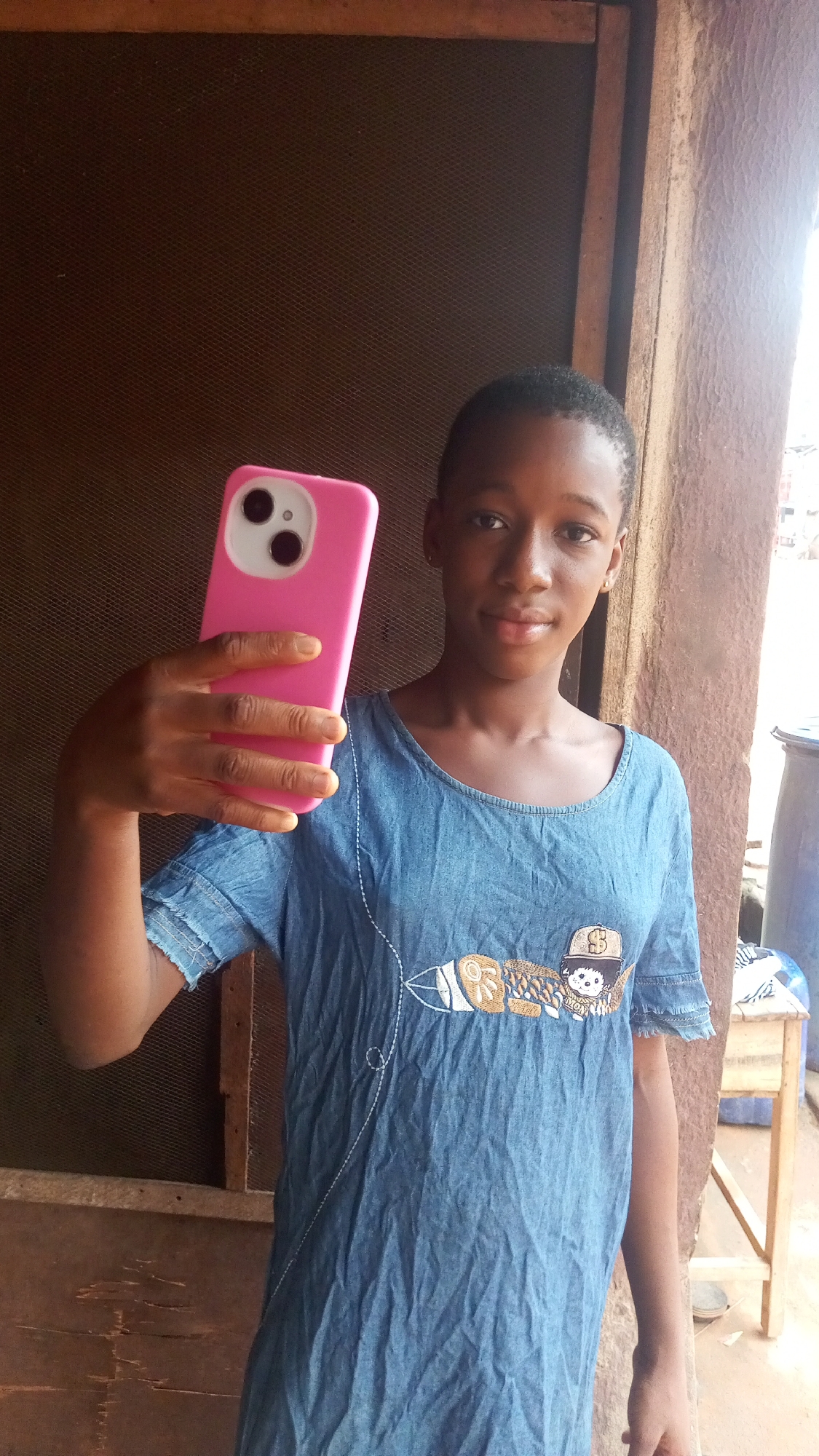A young woman stands indoors near a doorway or opening, holding a pink smartphone up in a selfie pose. She is wearing a denim-like shirt with a design embroidered or printed on the front. The background includes a rough, possibly unfinished wall or structure, and a glimpse of what appears to be an urban environment with other buildings in the distance. Based on the prompt, the location is Onitsha, Nigeria. Details like the type of construction, the woman's clothing, and the smartphone suggest a low-to-middle-income setting.