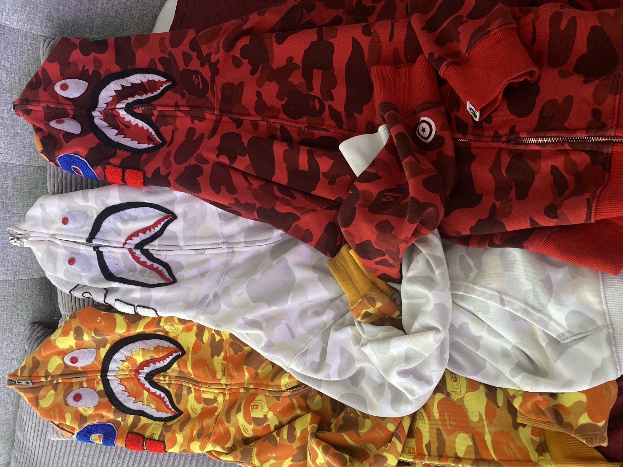 The image shows three Bathing Ape (BAPE) shark hoodies stacked on top of each other. The top hoodie is red camouflage, the middle hoodie is white camouflage, and the bottom hoodie is yellow camouflage. The hoodies are zippered and have the signature shark face graphic on the hood. The background appears to be a textured grey surface, possibly a couch or blanket. The image does not contain information about the location in the city of Sochi, Russia.