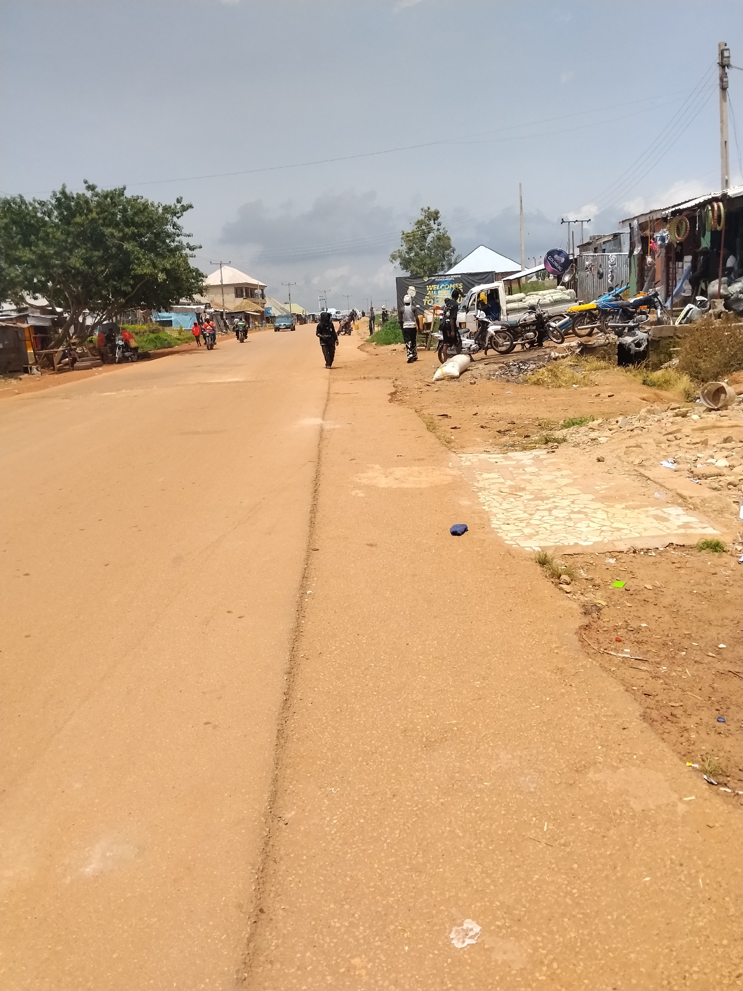 This image depicts a dusty, outdoor scene on a road in Durumi, Nigeria. The road, paved with reddish-brown asphalt, occupies the majority of the frame, with a dirt shoulder on the right. Several people are visible, including a person walking with a backpack, and others on or near motorcycles. A white truck, also with people around it, is parked on the right side of the road, accompanied by various motorcycles. On the far right, a row of open-air shops displays goods, including what appear to be tires, tools, and other merchandise. A large banner is visible in the background, with text that includes "WELCOME TO" and "DURUMI." The weather appears to be overcast, with a cloudy sky. The overall impression is that of a busy, commercial area on a typical day, likely in the morning or afternoon given the lighting.