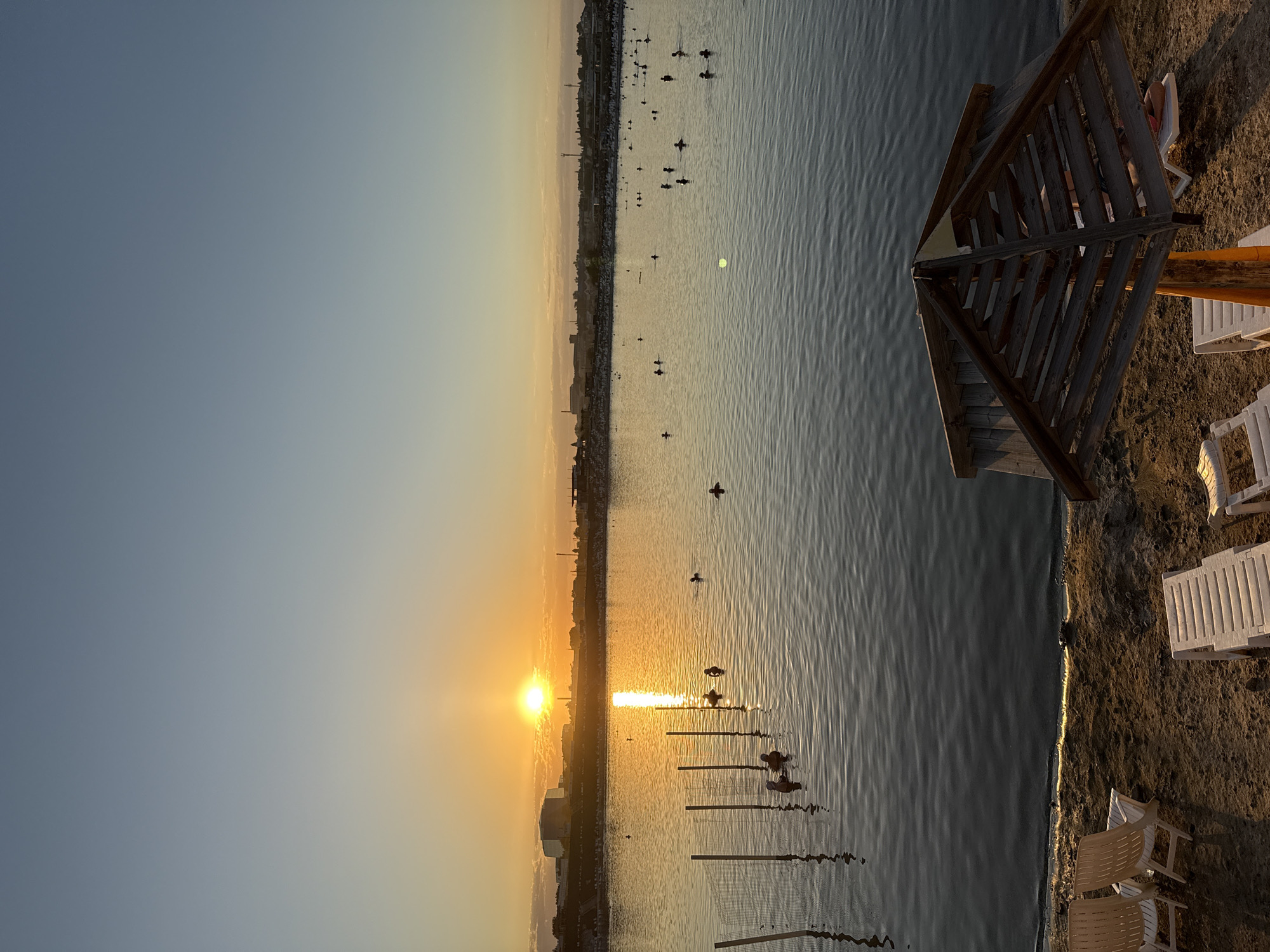 This media file captures a serene sunset over a body of water in Sol-Iletsk, Russia. The scene is dominated by the calm water, reflecting the warm hues of the setting sun. On the right side of the frame, a sandy beach is visible with several white lounge chairs and a wooden umbrella structure, indicating a recreational or resort area. Some people are seen relaxing on the chairs.
The activity taking place appears to be people enjoying the evening by the water. There are several individuals scattered across the water, some of whom appear to be on small watercraft or floating devices, while others might be swimming. The sun is low on the horizon, casting a bright, golden glow across the water and sky, suggesting it is either sunrise or sunset. The sky transitions from a clear, pale blue to soft orange and yellow near the sun. The water exhibits gentle ripples, adding to the tranquil atmosphere.
Notable details include the prominent sun low in the sky, creating a strong reflection on the water. The presence of beach furniture and the shoreline suggest a developed area for leisure. The overall mood is peaceful and relaxing, indicative of a beautiful evening by the water. There is no visible text in the image.