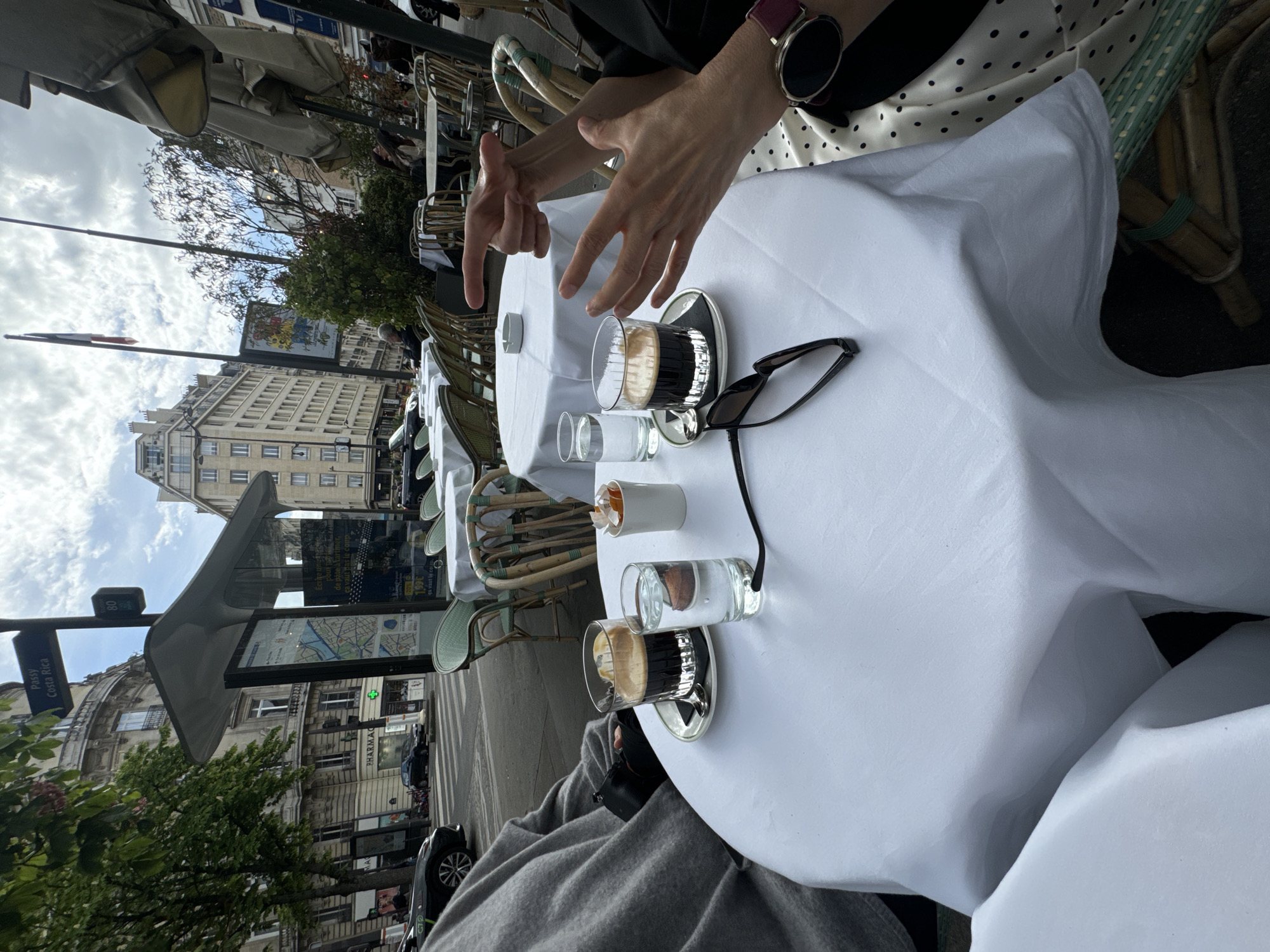 An overhead, slightly angled shot captures an outdoor cafe scene in what appears to be Paris, France. The image is rotated counter-clockwise.
At the center, two individuals are seated at a cafe table draped with a white tablecloth. Two cups of espresso, each on a saucer, and two small glasses of water are on the table. A pair of black sunglasses rests next to one of the coffee cups. A small white container, possibly for sugar or a candle, is also present. One person's hands are visible, gesturing expressively as if in conversation, wearing a watch on the left wrist and a white polka-dotted top. The other person's leg, clad in grey trousers, is visible, holding a black object (possibly a camera or phone) under the table.
The cafe features traditional wicker chairs with light green elements and large grey awnings or umbrellas providing shade. In the background, typical Parisian Haussmannian buildings line the street. A French flag flies on a pole in the distance. Street-side elements include trees, parked cars, and a bus stop shelter with a displayed map and advertisement.
The time of day is daytime, with a moderately cloudy sky. Visible text includes "Pansy" and "Costa Rica" on a street sign or informational panel, along with the number "80" and "0/0". A partial "PHARMAC" sign (likely "PHARMACIE") is visible on one of the buildings. Text on the bus stop advertisement includes partial French words like "Escapade" and numbers like "150€". The overall scene depicts a relaxed, social interaction at an outdoor cafe in a bustling city environment.