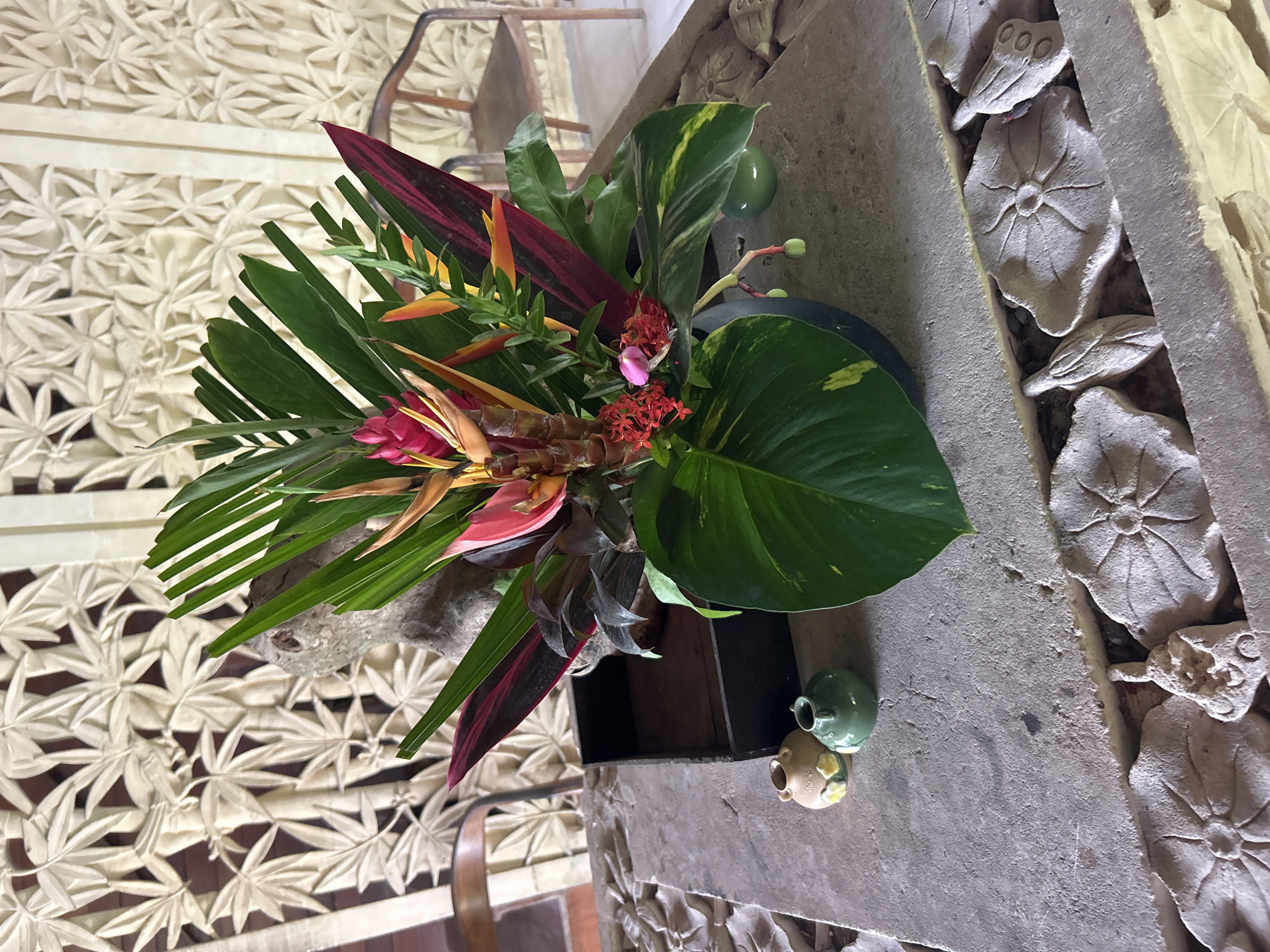 A tropical flower arrangement is displayed on a stone table in a room with intricately carved walls and wooden chairs. The arrangement features vibrant red, orange, and yellow heliconia flowers, alongside large green leaves and dark red foliage. Two small ceramic vases, one tan with yellow floral accents and the other green, are placed to the left of the arrangement. The carved walls behind the table have a repetitive pattern of stylized leaves and branches. The floor appears to be made of textured stone. The scene suggests an interior space in a tropical setting, possibly a hotel or a traditional Balinese building in Mengwi, Indonesia.